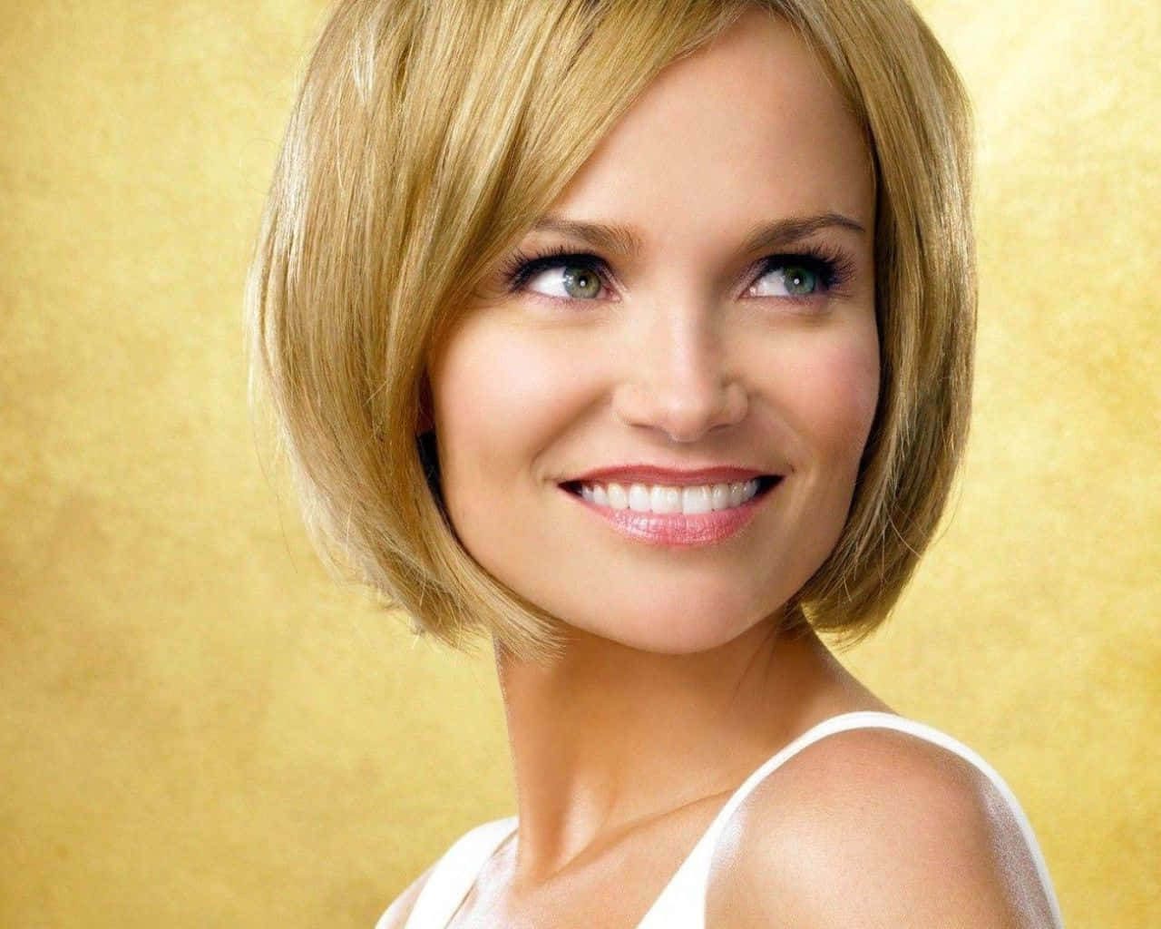 kristin chenoweth 桌布 壁紙 — 一位留著金色鮑伯頭、笑容燦爛的女性，在溫暖的金色紋理背景上向上凝視。