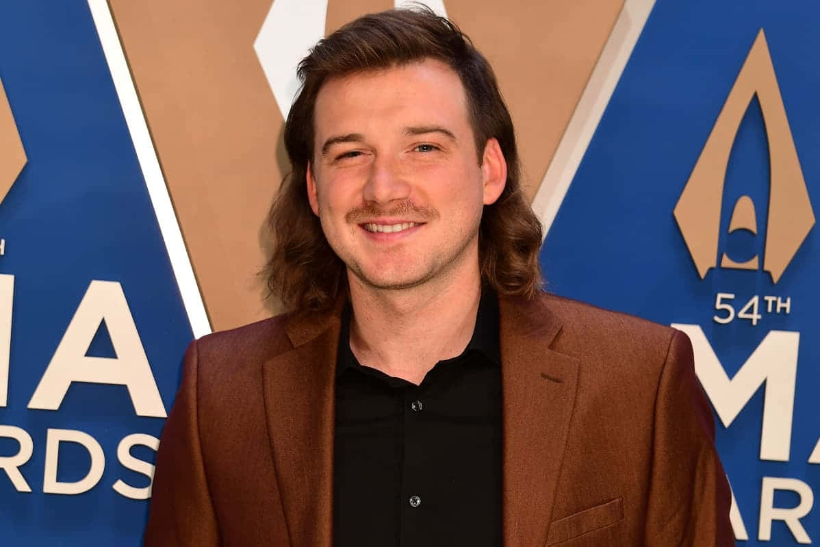 Morgan Wallen 桌布 壁紙 — 一位留著長棕髮和鬍鬚的微笑男士，在第54屆AMA頒獎典禮上穿著棕色西裝外套，內搭黑色襯衫。
