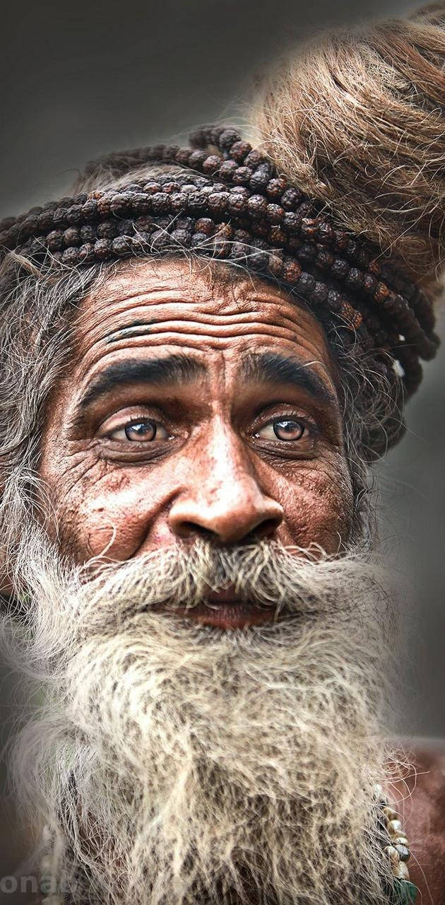 Aghori 桌布 壁紙 — 一位留著長灰鬍鬚和辮子頭髮並飾有珠子的老人的特寫肖像。
