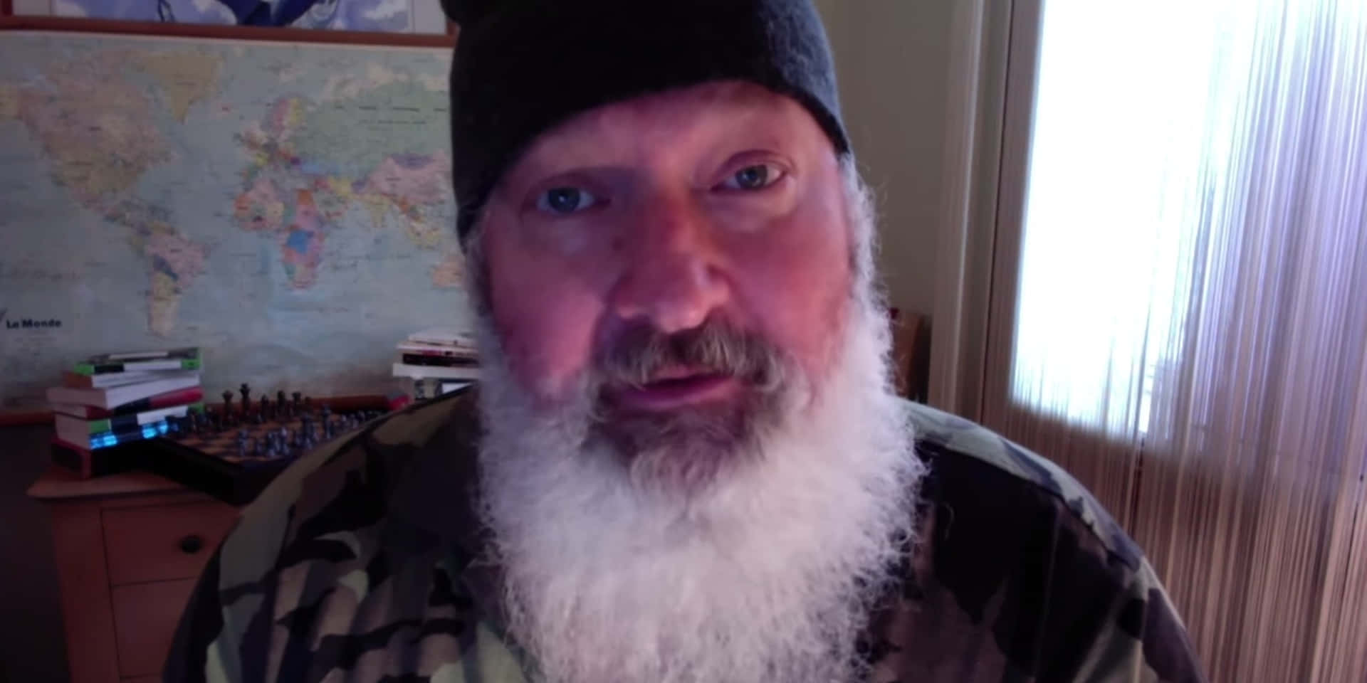 Randy Quaid 桌布 壁紙 — 一位留著長長白鬍子、戴著無邊便帽的男人，在一個有世界地圖和棋盤的房間裡直視鏡頭。