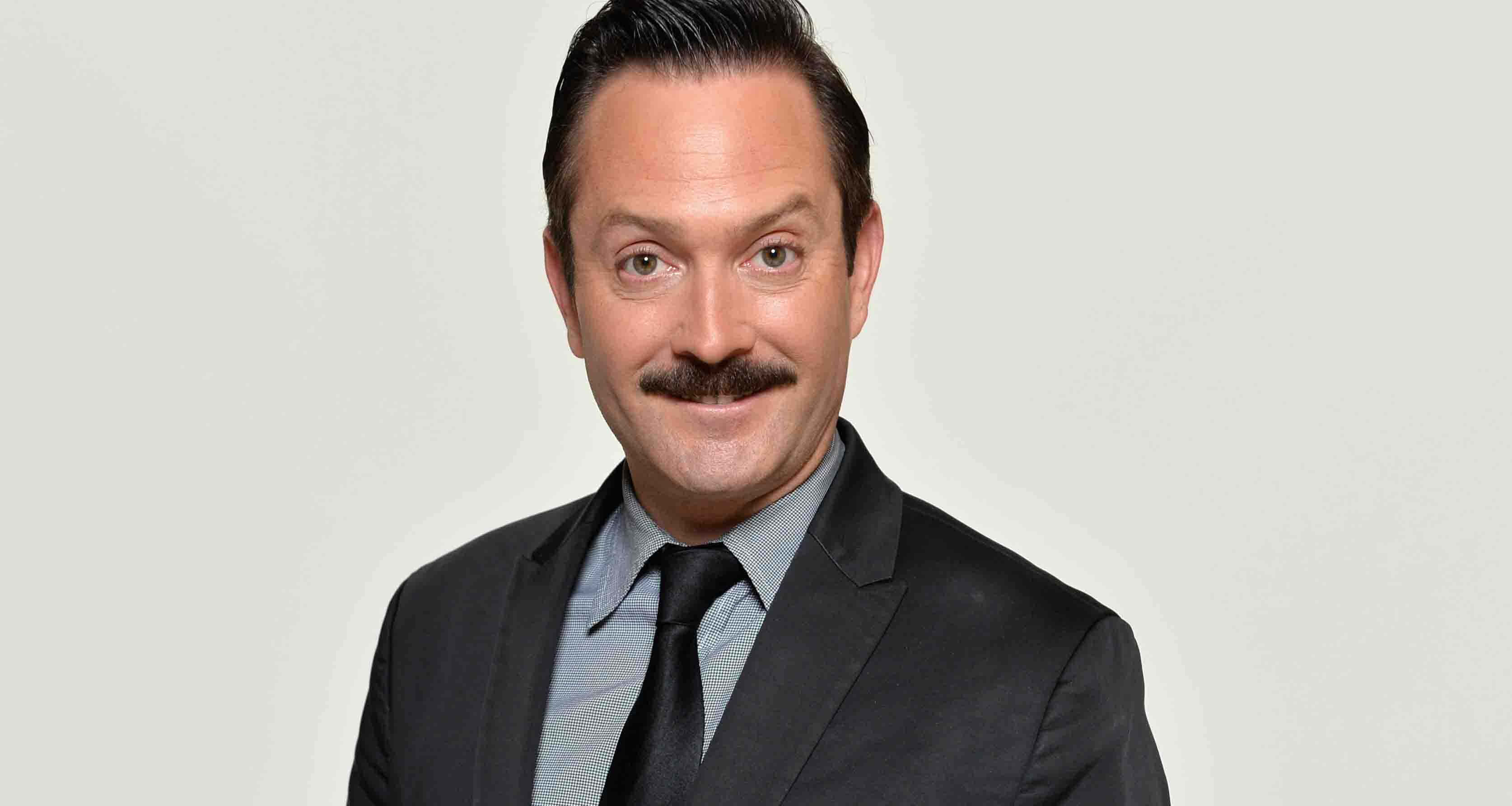 thomas lennon 桌布 壁紙 — 一位留著顯眼鬍鬚的男人，身穿深色西裝，在純淺灰色背景前自信地微笑。