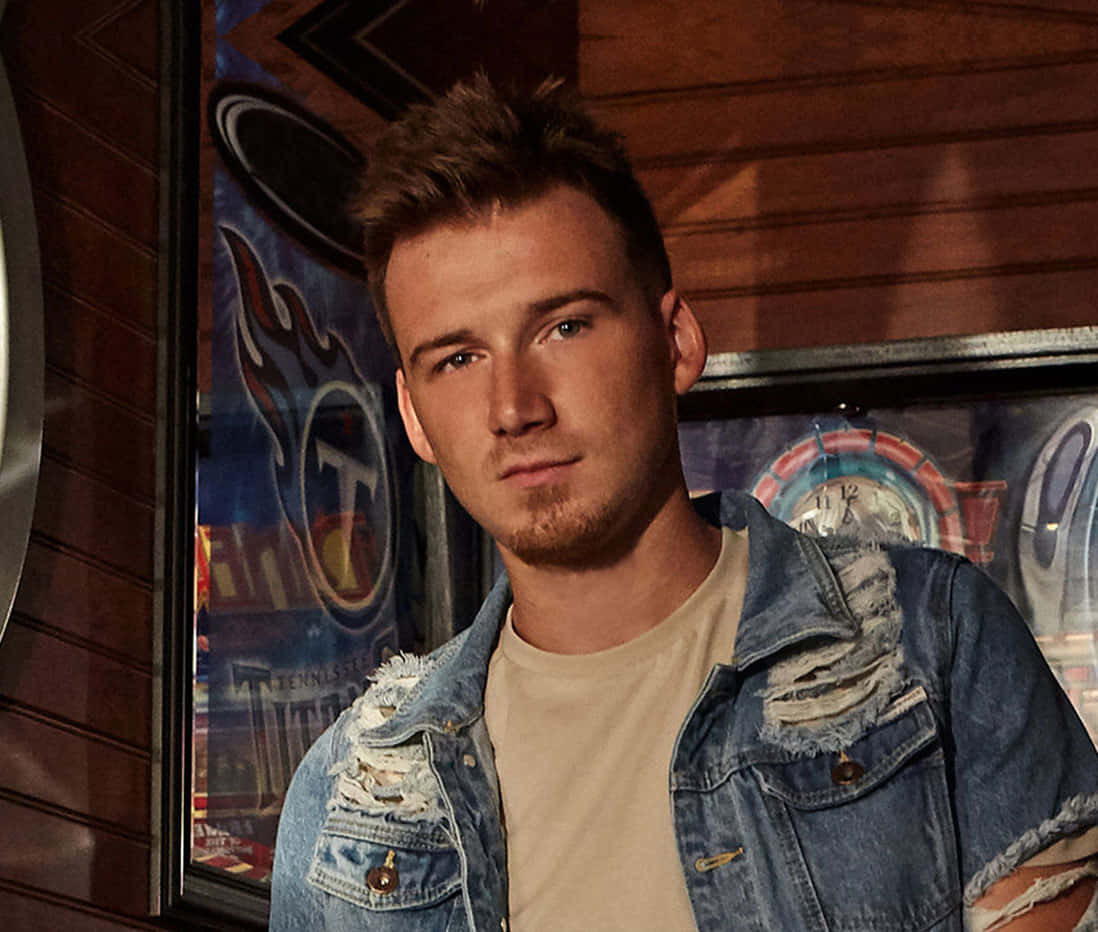 Morgan Wallen 桌布 壁紙 — 一位留著鬍渣和刺蝟頭的年輕男子，身穿破洞牛仔外套內搭米色T恤，站在光線昏暗的酒吧裡，身後是木牆和飛鏢靶海報。