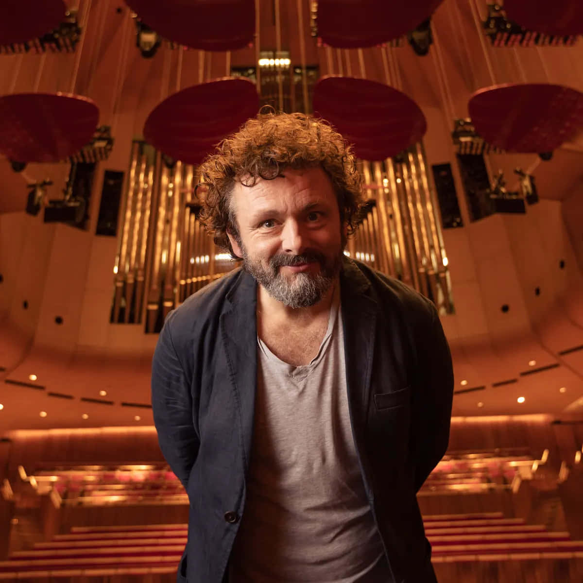 Michael Sheen 桌布 壁紙 — 一位留著鬍鬚、捲髮的男子在一個有發光管和紅色座位的宏偉音樂廳中溫暖地微笑。