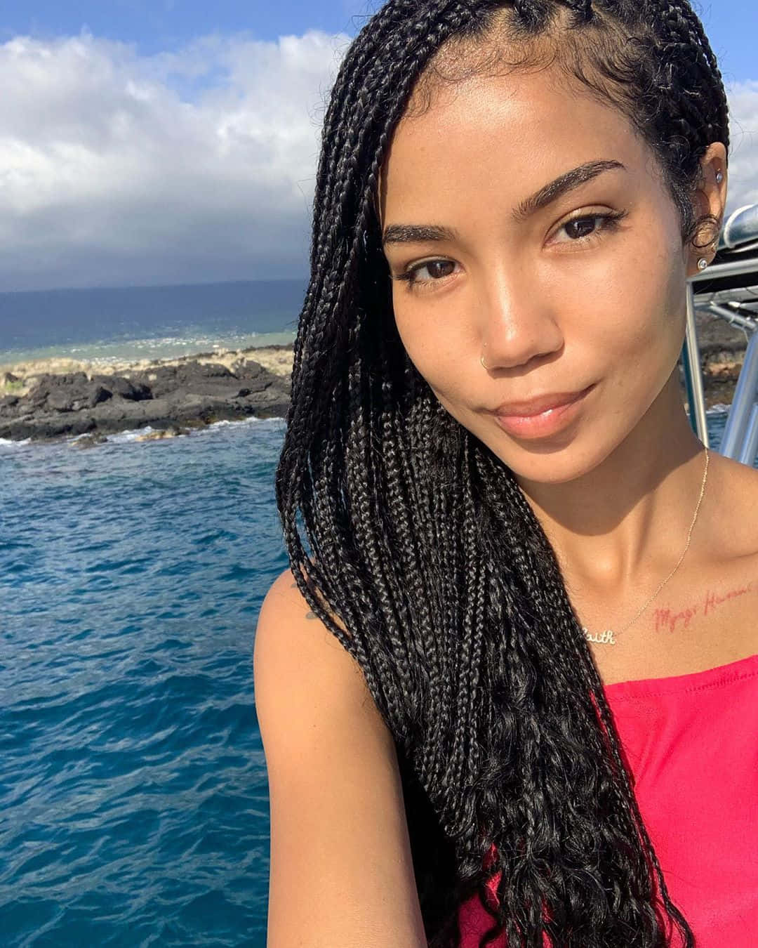 jhene aiko 桌布 壁紙 — 一位留著黑色長辮子的女性在船上微笑，背景是海洋和岩石海岸，天空部分多雲。