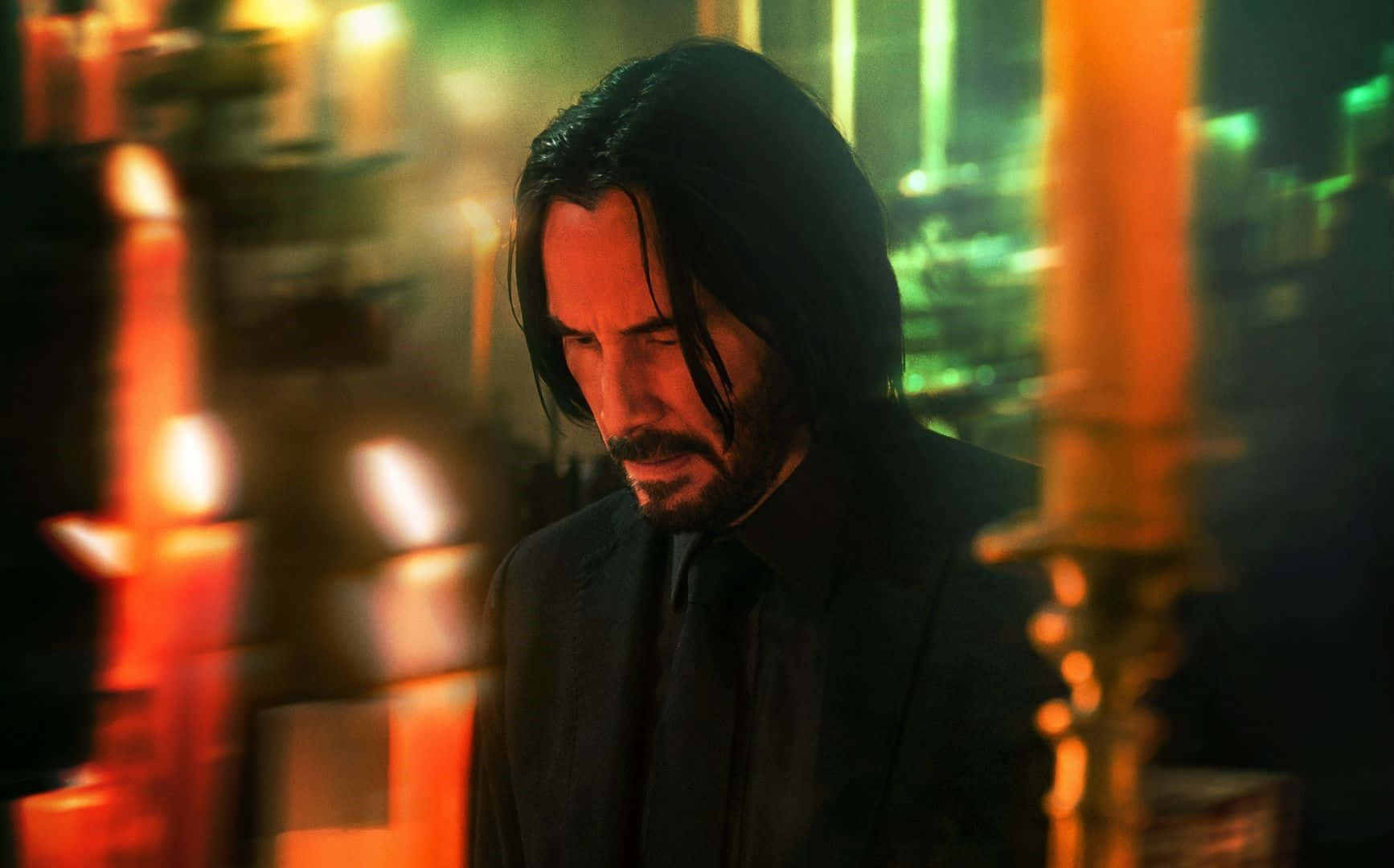 Keanu Reeves 桌布 壁紙 — 一位留著黑色長髮和鬍鬚、身穿黑色西裝的男子，莊嚴地站在一個光線昏暗的房間裡，背景是模糊的橙色和綠色燭光。
