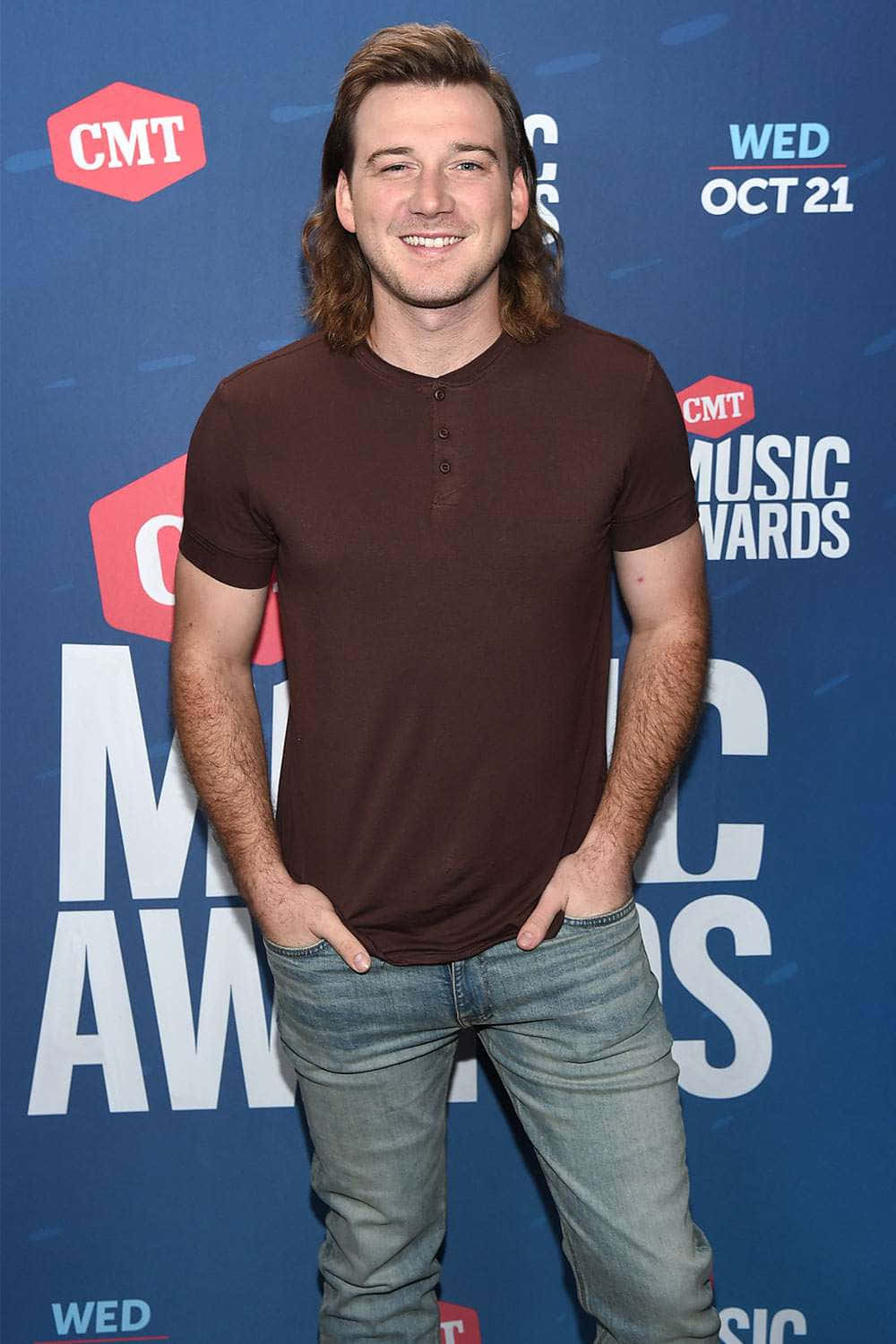 Morgan Wallen 桌布 壁紙 — 一位留長髮、身穿棕色亨利衫和牛仔褲、面帶微笑的男子在CMT音樂獎紅地毯上自信地擺姿勢。