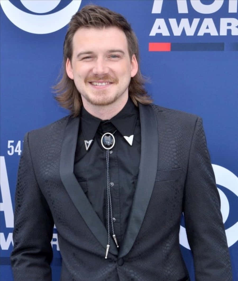 Morgan Wallen 桌布 壁紙 — 一位留長髮和鬍鬚的微笑男士，身穿黑色紋理西裝，配以波洛領帶，背景為藍色頒獎典禮。