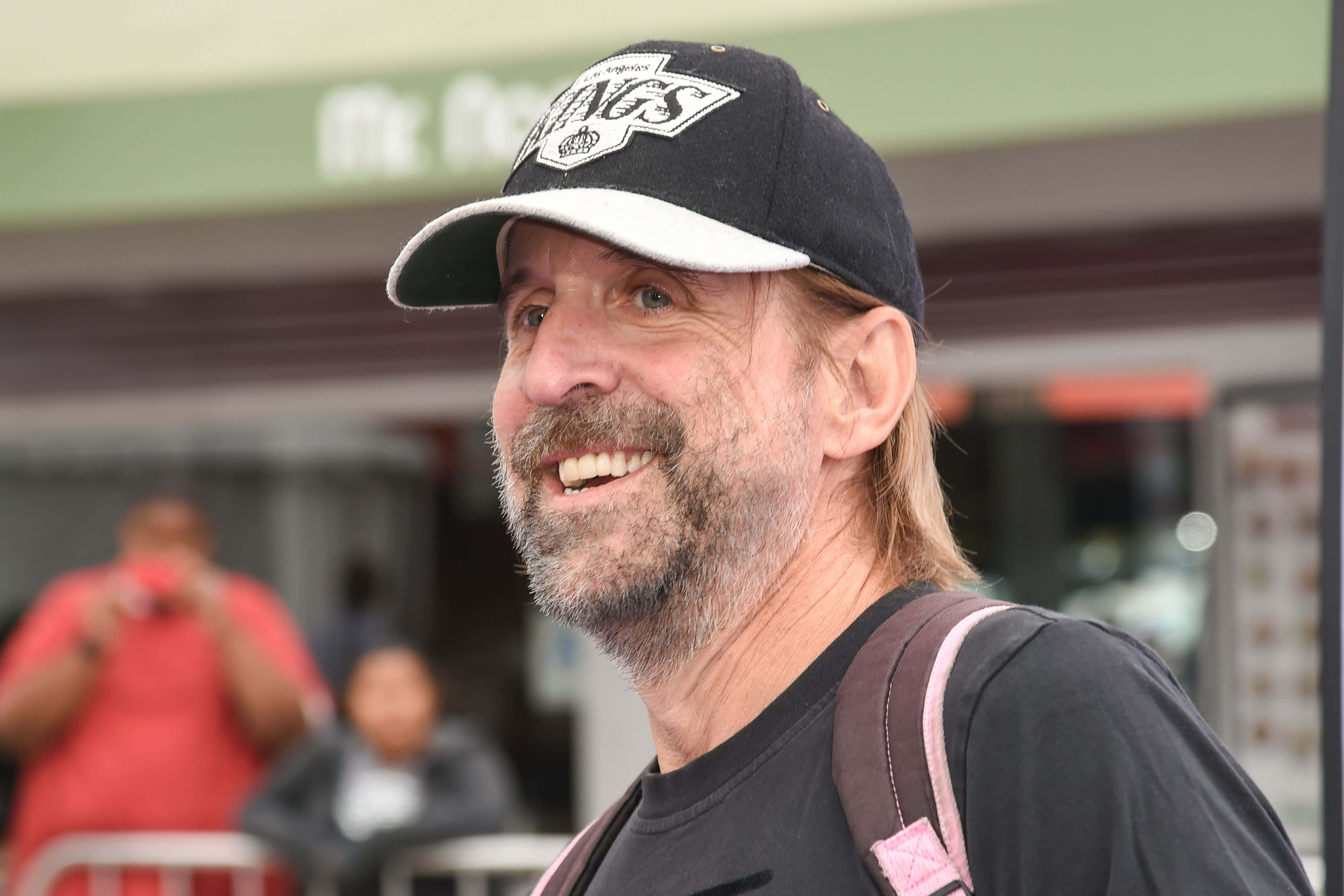 Peter Stormare 桌布 壁紙 — 一位留鬍子、戴帽子的開朗中年男子，揹著背包在戶外溫暖地微笑。