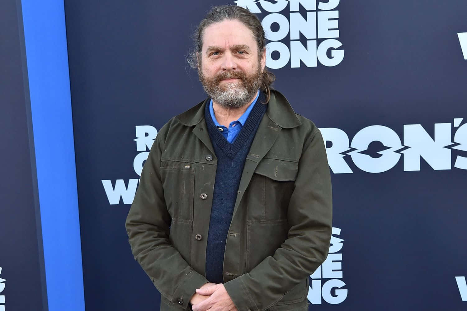 Zach Galifianakis 桌布 壁紙 — 一位留鬍子、長髮的男子自信地站在藍色促銷背景前，背景上有白色文字。