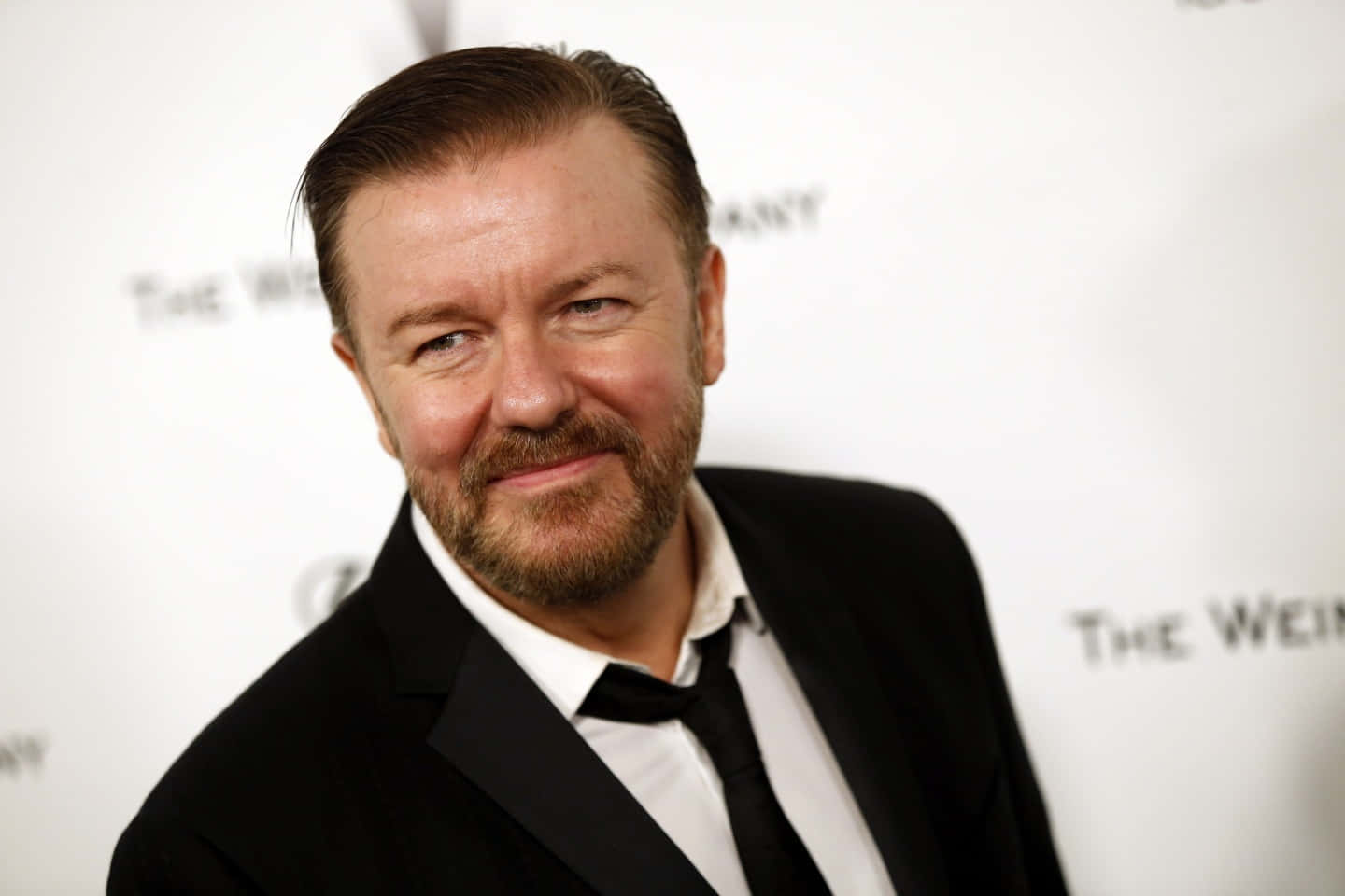 ricky gervais 桌布 壁紙 — 一位留鬍子、頭髮向後梳的男人，身穿黑色西裝，在白色品牌背景前微笑。