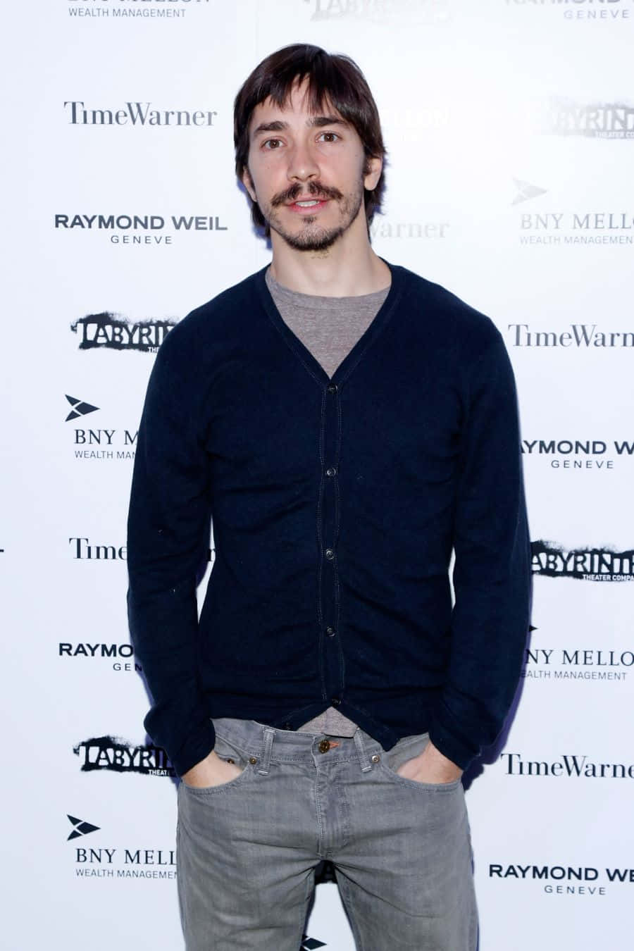 Justin Long 桌布 壁紙 — 一位留鬍子、黑髮的男子自信地站在記者會上，穿著海軍藍開襟衫和灰色牛仔褲，背景是品牌背景。