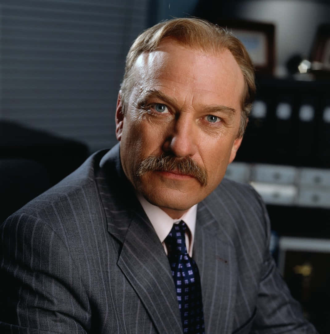 Ted Levine 桌布 壁紙 — 一位留鬍子的嚴肅男子穿著細條紋西裝，在燈光昏暗的辦公室裡專注地凝視著相機。