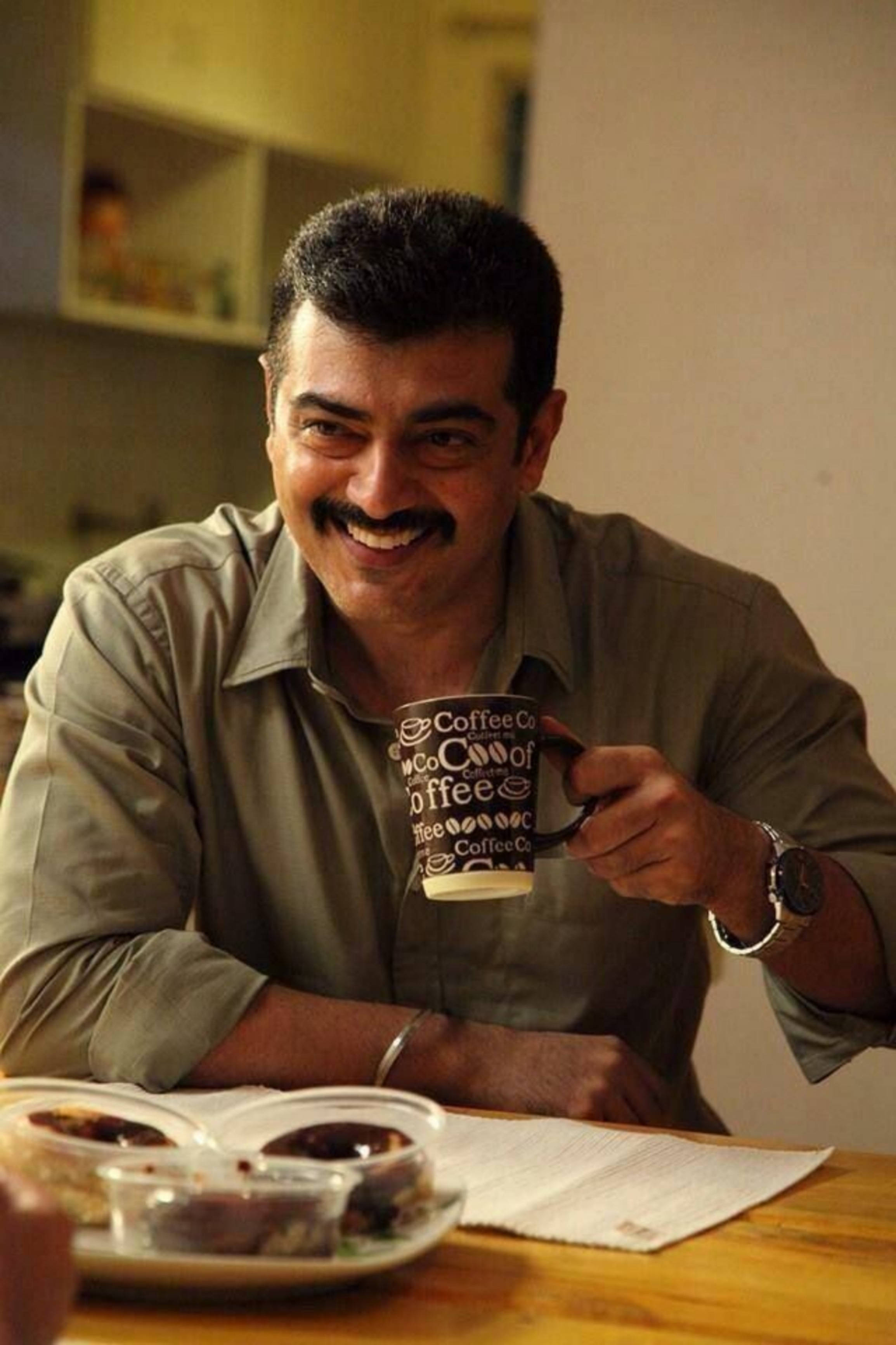 Tamil Actors HD 桌布 壁紙 — 一位留鬍子的微笑男士在木桌上拿著咖啡杯，旁邊有糕點和文件。