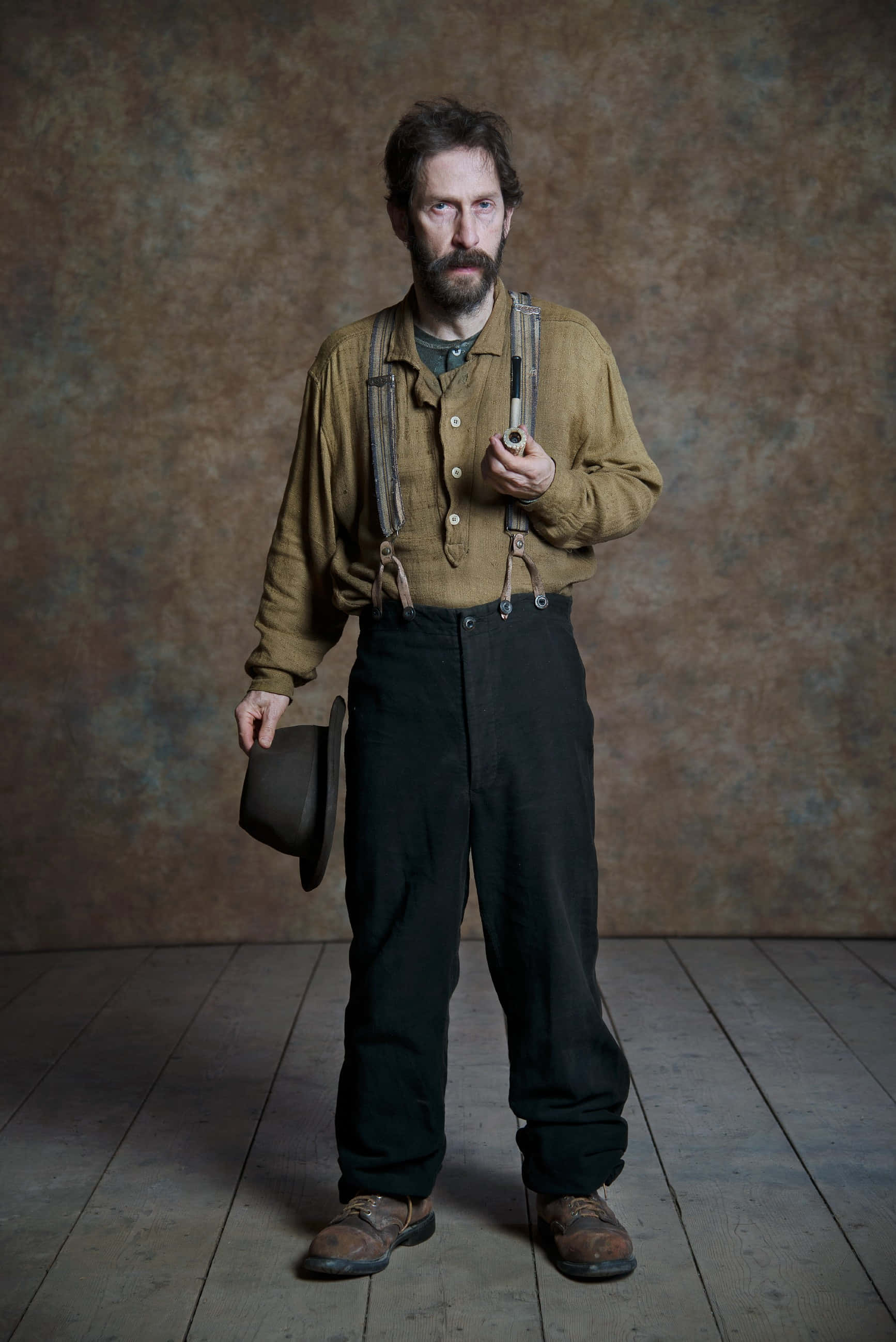 Tim Blake Nelson 桌布 壁紙 — 一位留鬍子的男士穿著復古服裝，莊嚴地拿著煙斗和帽子，背景有紋理。