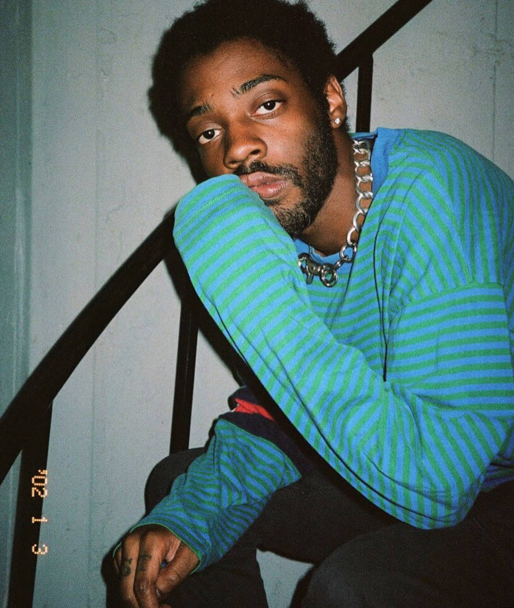 Brent Faiyaz 桌布 壁紙 — 一位留鬍鬚、戴鏈條項鍊的年輕男子穿著藍綠條紋毛衣坐在樓梯上，直視鏡頭。