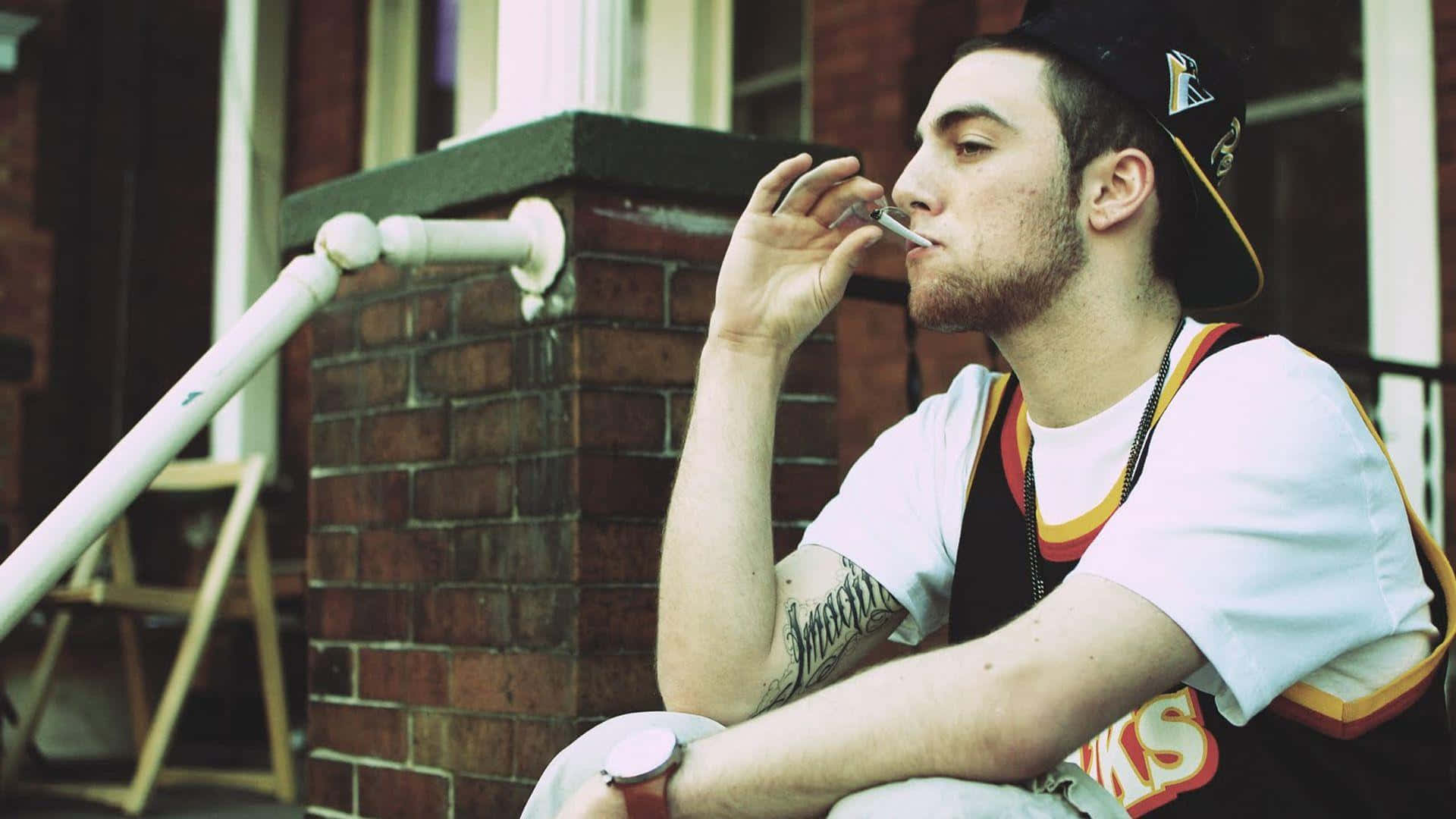 Mac Miller 桌布 壁紙 — 一位留鬍鬚、手臂有紋身的年輕男子坐在磚砌建築外的台階上吸煙。
