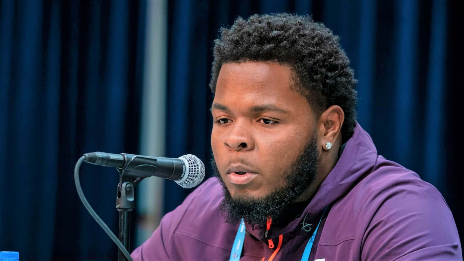 Jawaan Taylor 桌布 壁紙 — 一位留鬍鬚、捲髮的男子，穿著紫色連帽衫，在藍色窗簾背景前對著麥克風說話。