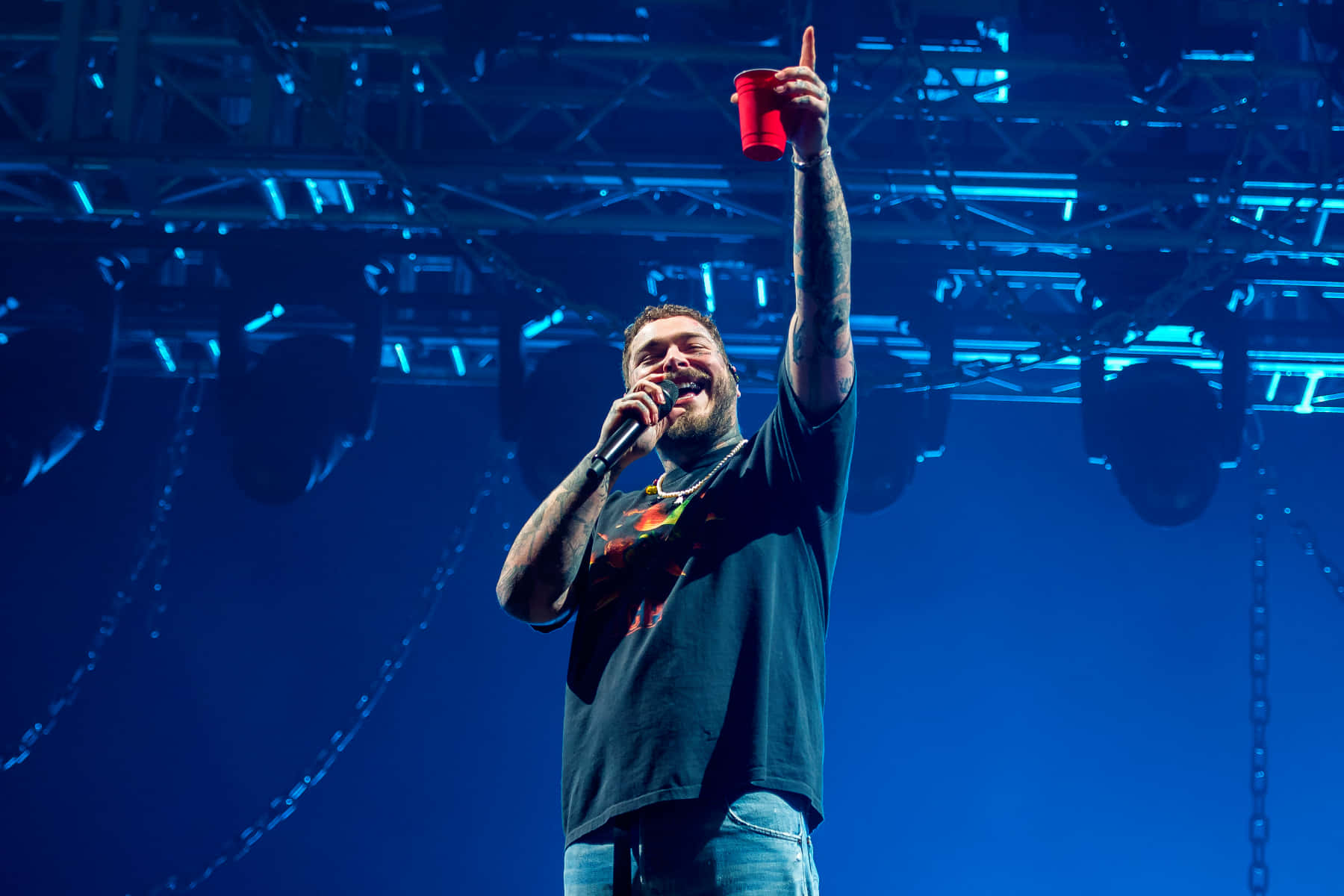 Post Malone 桌布 壁紙 — 一位留鬍鬚、有紋身的男士在藍色燈光的舞台上，一邊舉起紅色杯子，一邊對著咪高峰唱歌。