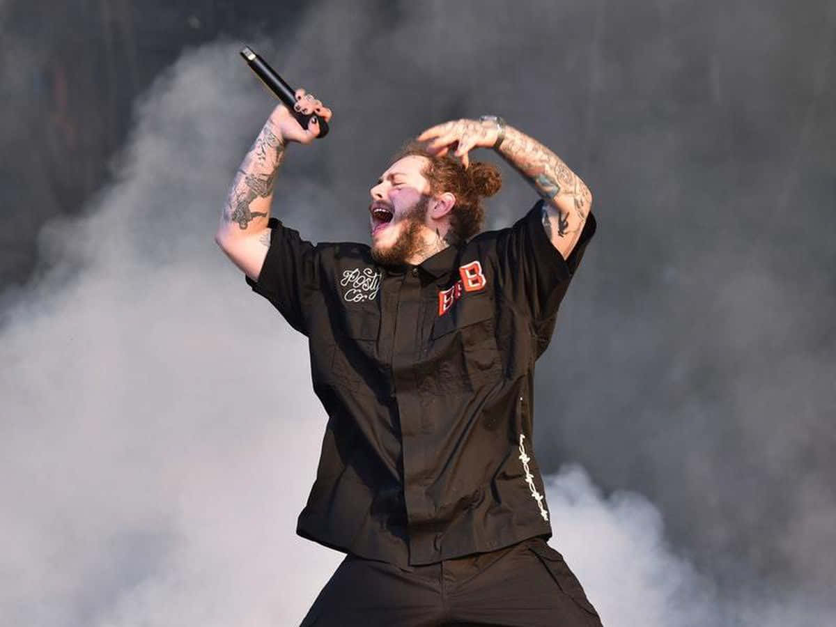 Post Malone 桌布 壁紙 — 一位留鬍鬚、有紋身的男歌手在煙霧繚繞中手持咪高峰，充滿激情地在舞台上表演。