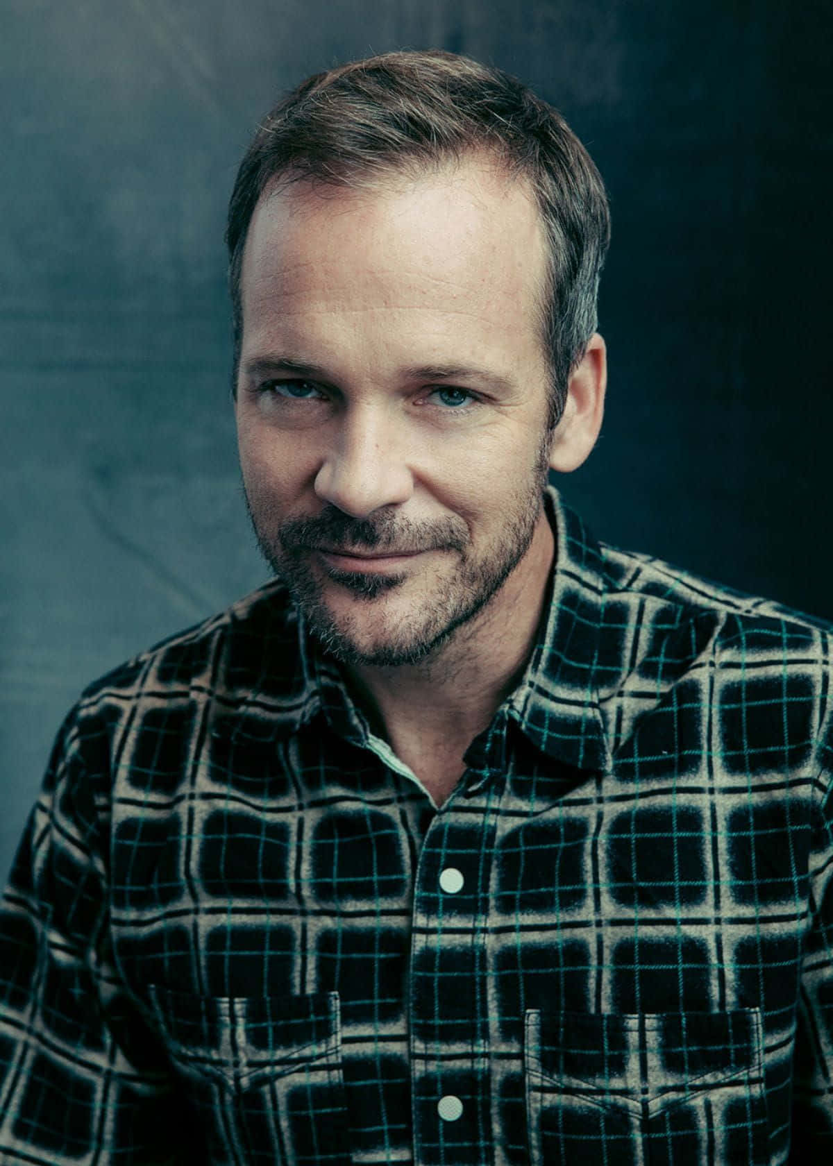 Peter Sarsgaard 桌布 壁紙 — 一位留鬍鬚、藍眼睛的男人，穿著格仔襯衫，在深色紋理背景前溫柔微笑。