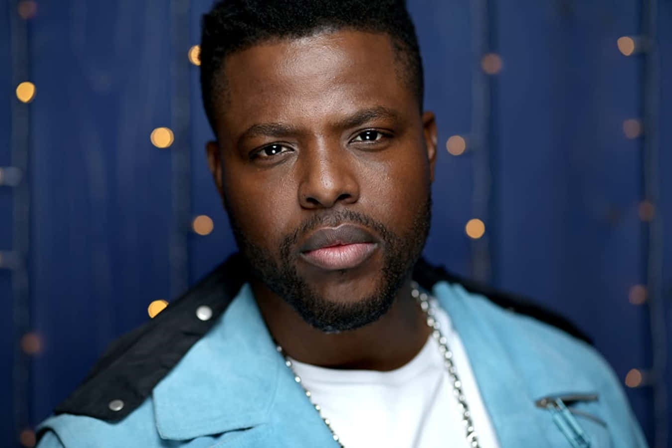 Winston Duke 桌布 壁紙 — 一位留鬍鬚、表情嚴肅的男子，身穿藍色外套，背景是帶有溫暖散景燈光的藍色背景。