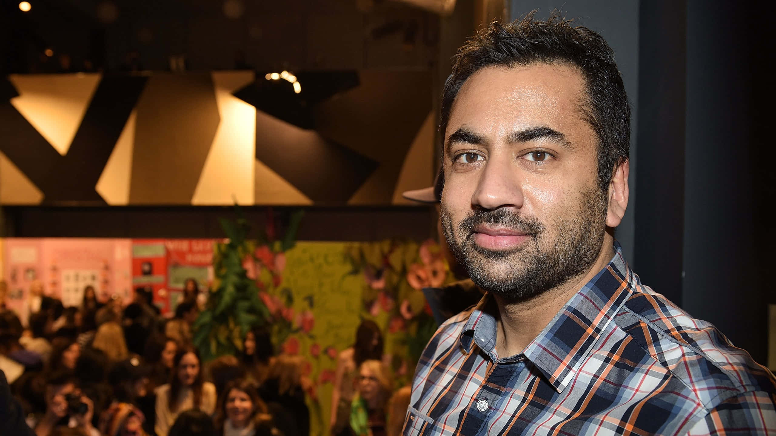 Kal Penn 桌布 壁紙 — 一位留鬍鬚、身穿格仔恤衫嘅男士喺燈光溫暖嘅室內活動中望向鏡頭，背後有模糊嘅人群同花卉裝飾。