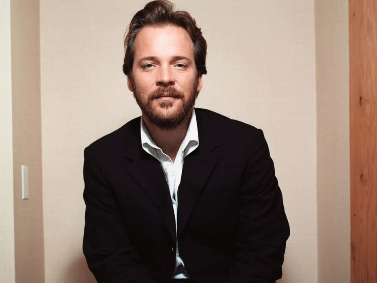 Peter Sarsgaard 桌布 壁紙 — 一位留鬍鬚、身穿深色西裝的男子，平靜地靠著帶有木飾邊的中性色牆壁坐著。