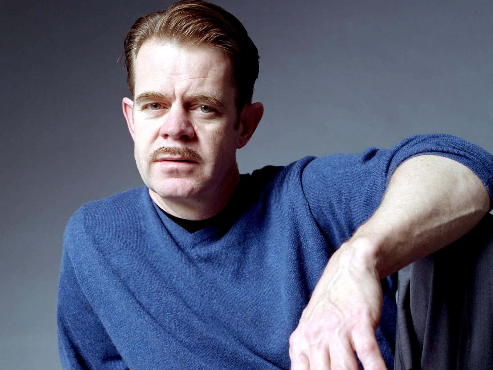 william h. macy 桌布 壁紙 — 一位留鬍鬚、身穿藍色毛衣的男子，在灰色背景前身體前傾，表情嚴肅。
