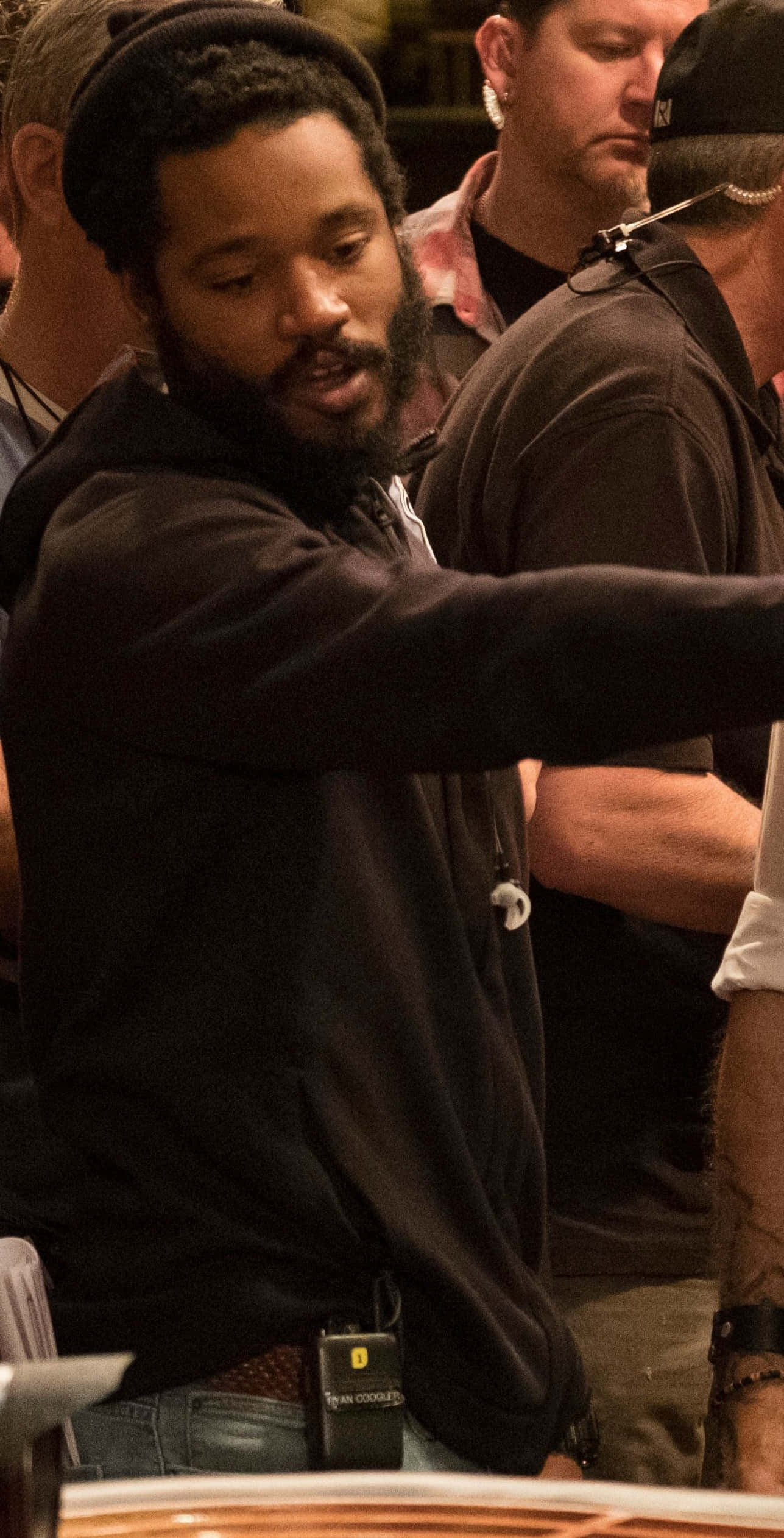 Ryan Coogler 桌布 壁紙 — 一位留鬍鬚戴冷帽的男子在擠滿人的室內環境中，在溫暖的燈光下生動地做手勢。