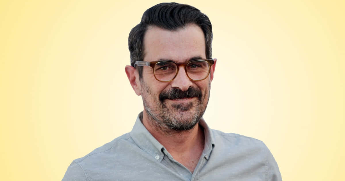 Ty Burrell 桌布 壁紙 — 一位留鬍鬚戴眼鏡的男子在柔和黃色背景前溫暖微笑。