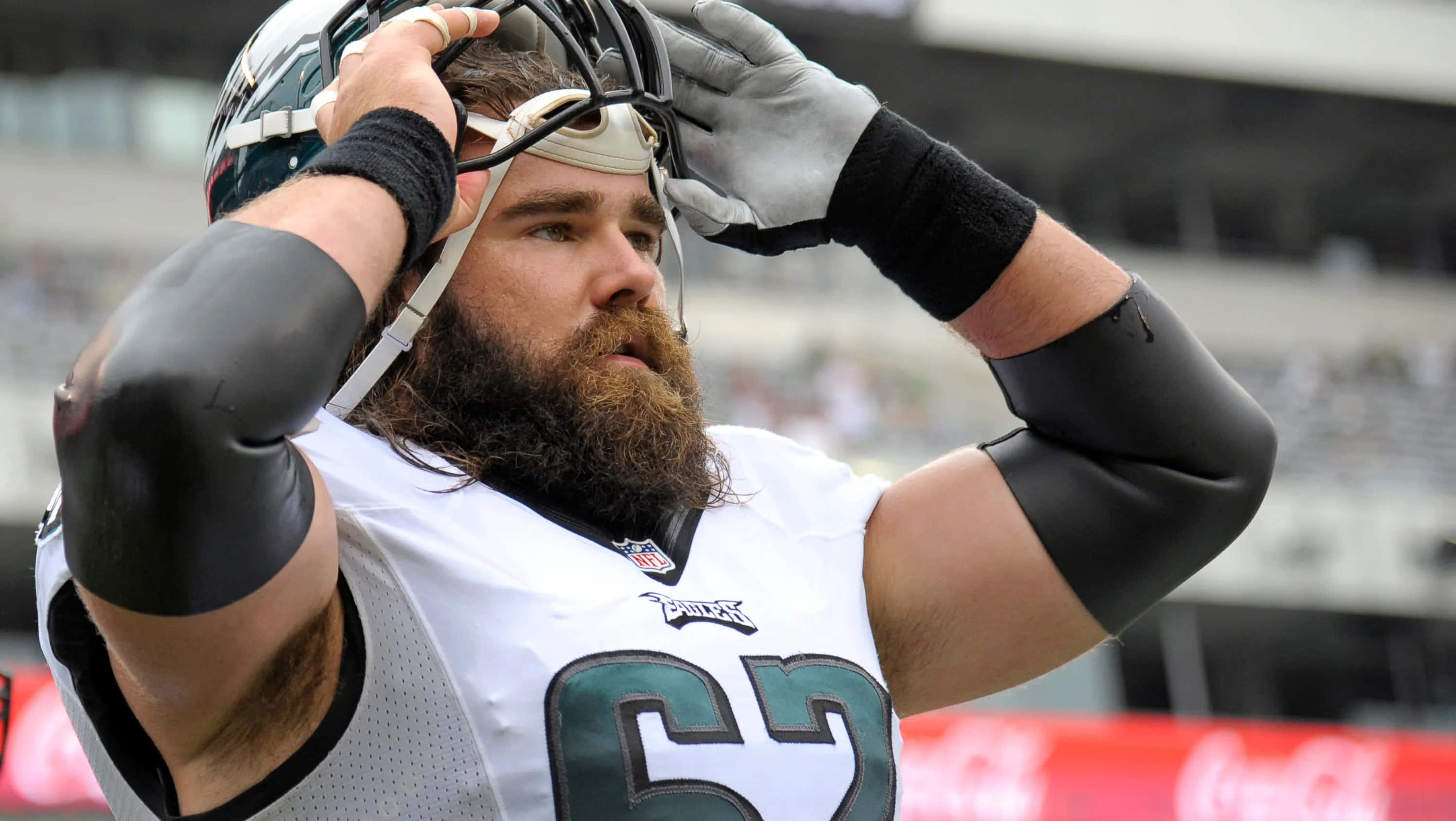 Jason Kelce 桌布 壁紙 — 一位留鬍鬚的NFL球員身穿白色老鷹隊球衣，在球場上專注地調整頭盔。