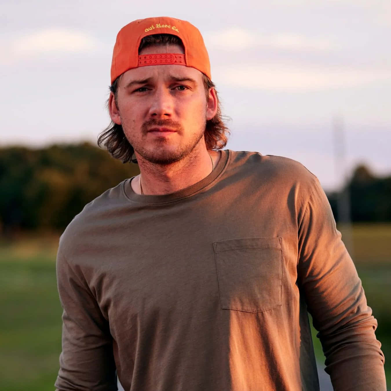 Morgan Wallen 桌布 壁紙 — 一位留鬍鬚的年輕男子戴著橙色帽子，在黃金時刻柔和溫暖的光線下站在田野中。