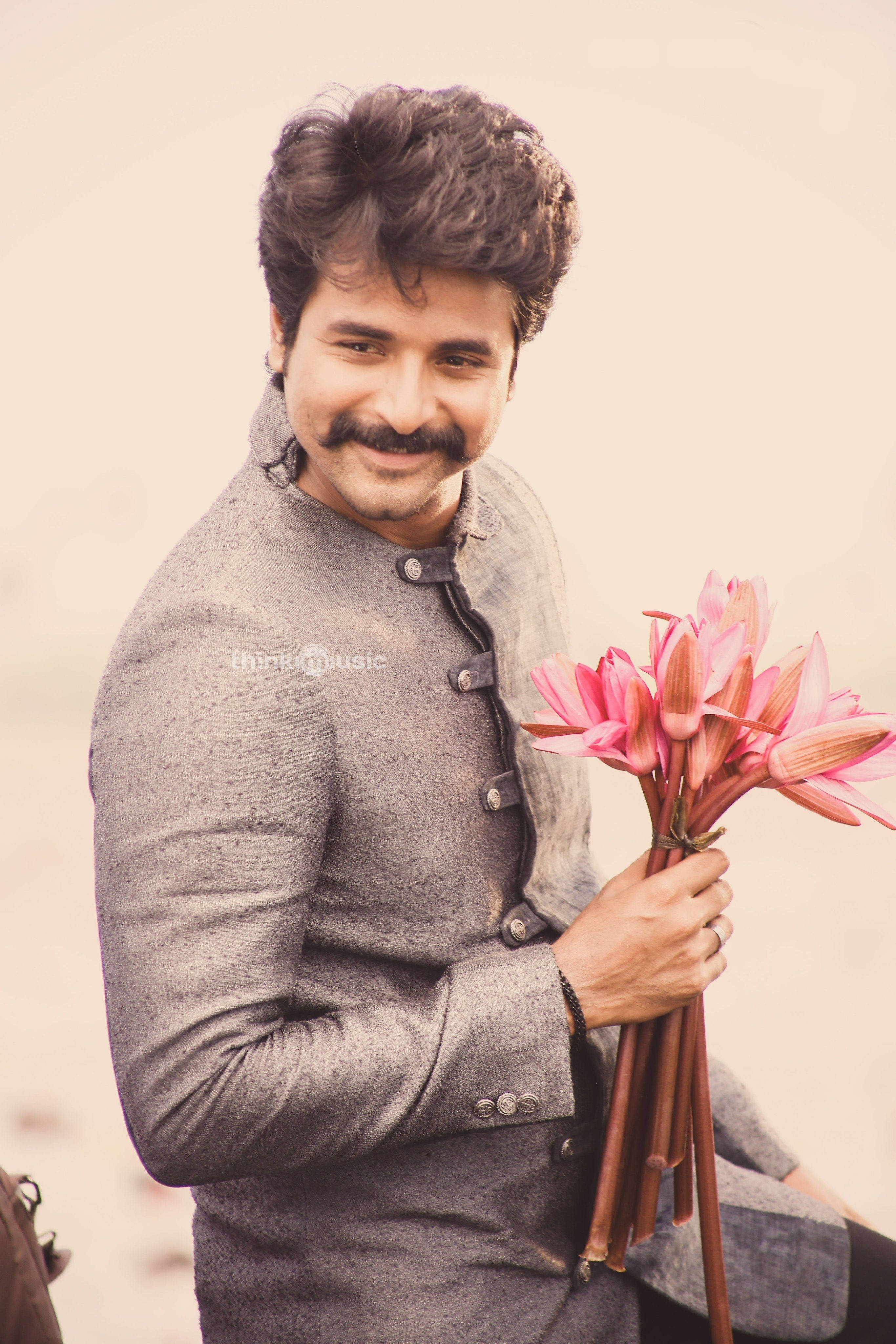 Sivakarthikeyan 桌布 壁紙 — 一位留鬍鬚的微笑男士，在柔和的米色背景前拿著一束粉紅色的花。