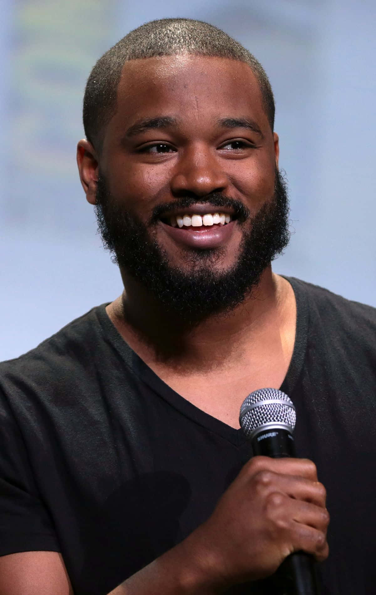 Ryan Coogler 桌布 壁紙 — 一位留鬍鬚的微笑男士在座談會活動中手持麥克風，身穿黑色T恤。