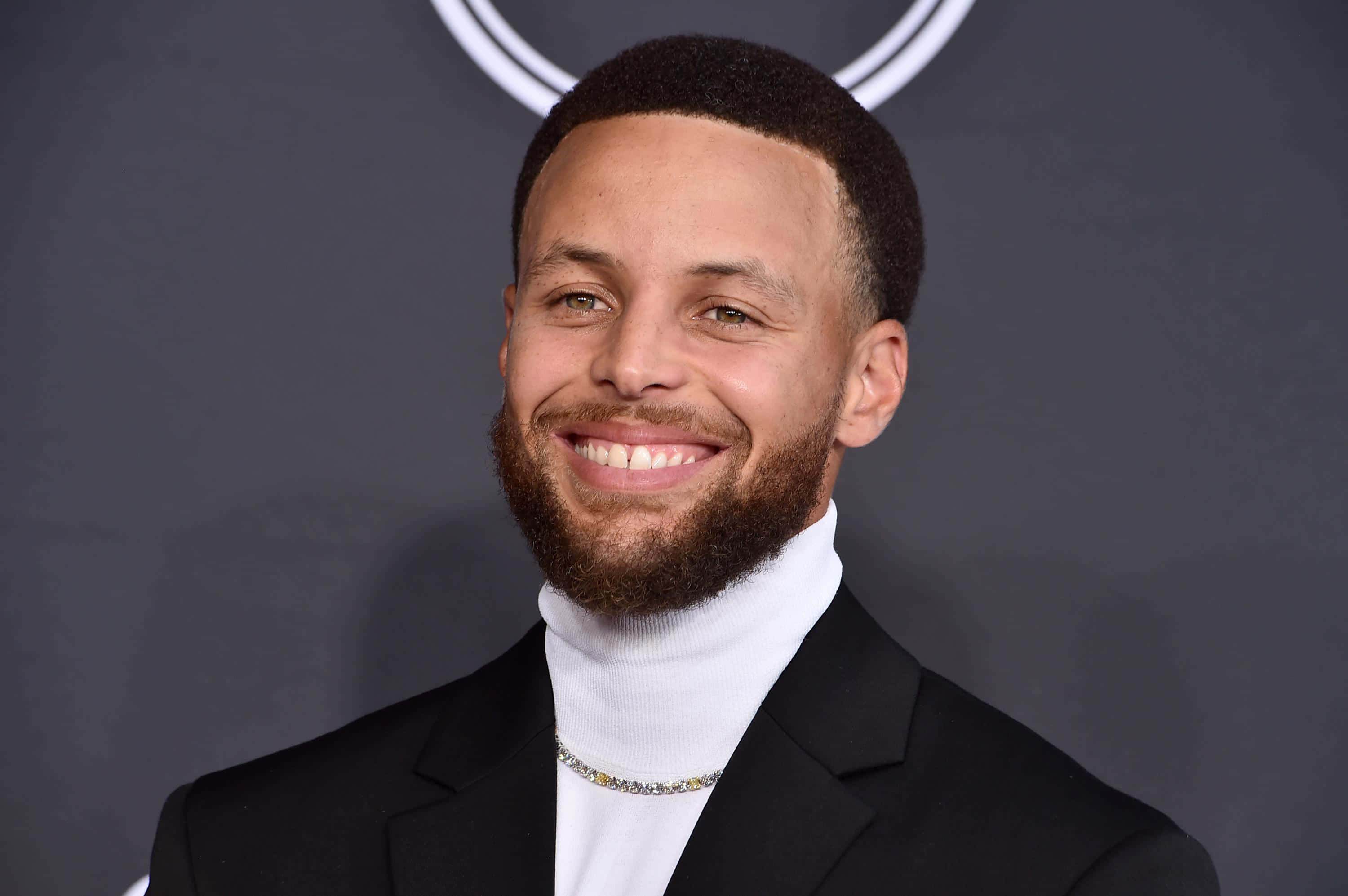 steph curry 桌布 壁紙 — 一位留鬍鬚的微笑男士穿著黑色西裝和白色高領毛衣，背景為深灰色並帶有圓形標誌。