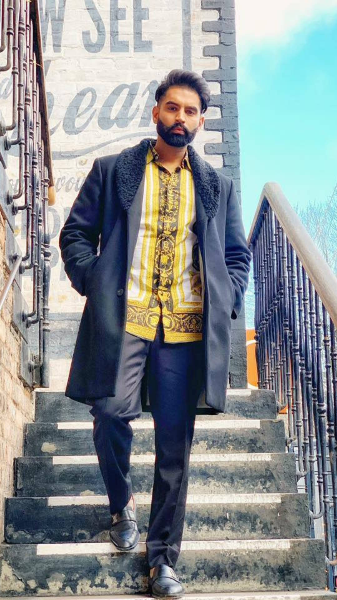 Indian 桌布 壁紙 — 一位留鬍鬚的時尚男士自信地站在風化的石階上，身穿深色外套，內搭大膽的黃色圖案襯衫。