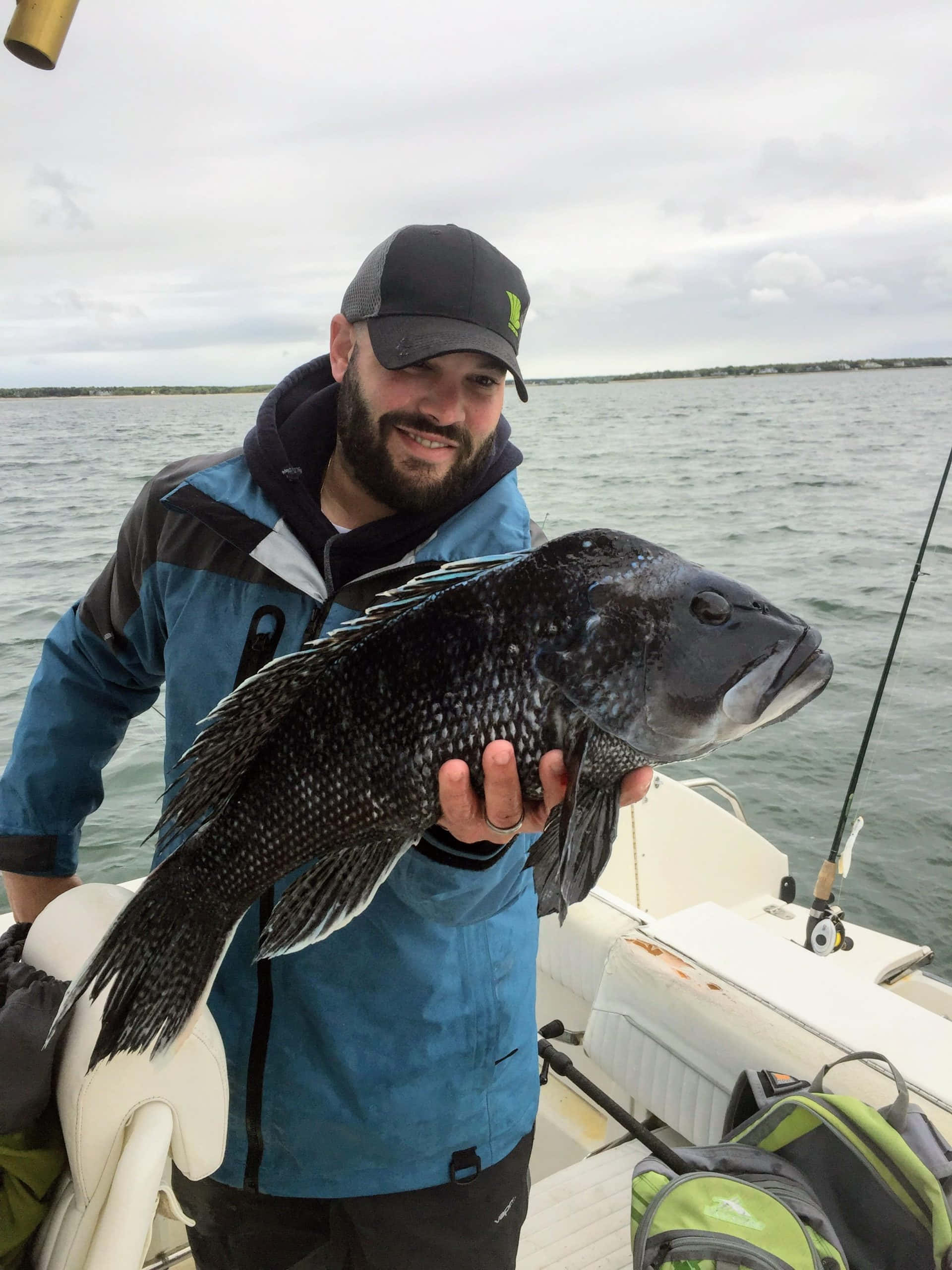 Black Sea Bass 桌布 壁紙 — 一位留鬍鬚的漁夫穿著藍色外套，在平靜的水面和陰天背景下，自豪地在船上拿著一條有斑點的大魚。