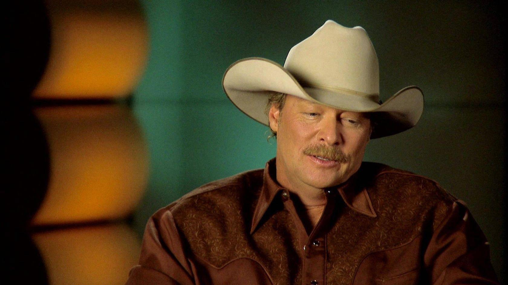 Alan Jackson 桌布 壁紙 — 一位留鬍鬚的男人，戴著白色牛仔帽和穿著棕色襯衫，在昏暗的房間裡向下看，身後有溫暖模糊的燈光。