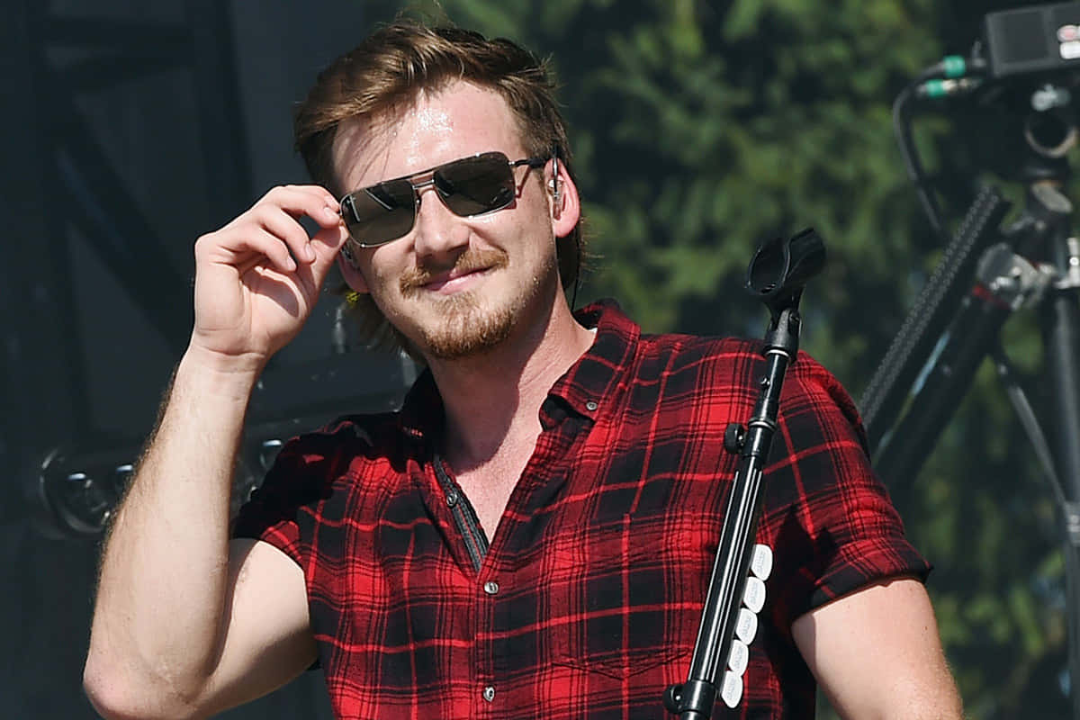 Morgan Wallen 桌布 壁紙 — 一位留鬍鬚的男士面帶微笑，在陽光明媚的戶外舞台上穿著紅色格仔襯衫，調整他的太陽眼鏡。