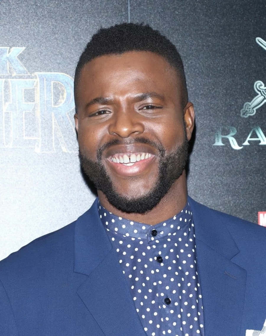 Winston Duke 桌布 壁紙 — 一位留鬍鬚的男士面帶微笑，身穿藍色西裝和圓點襯衫，背景是深色活動場景。