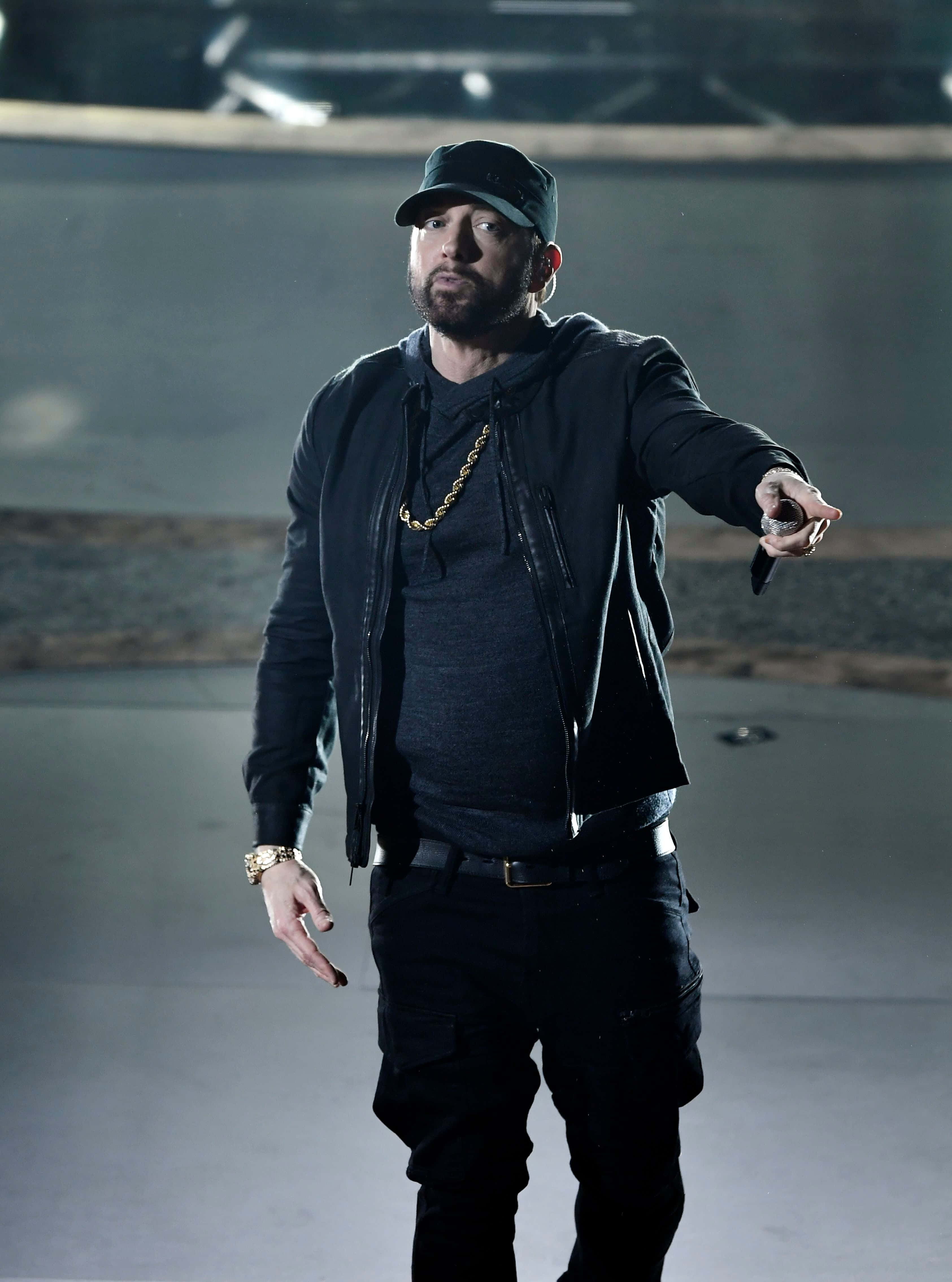 Eminem 桌布 壁紙 — 一位留鬍鬚的男子戴著黑色帽子和外套，在昏暗的工業環境中，於戲劇性的舞台燈光下向前做手勢。