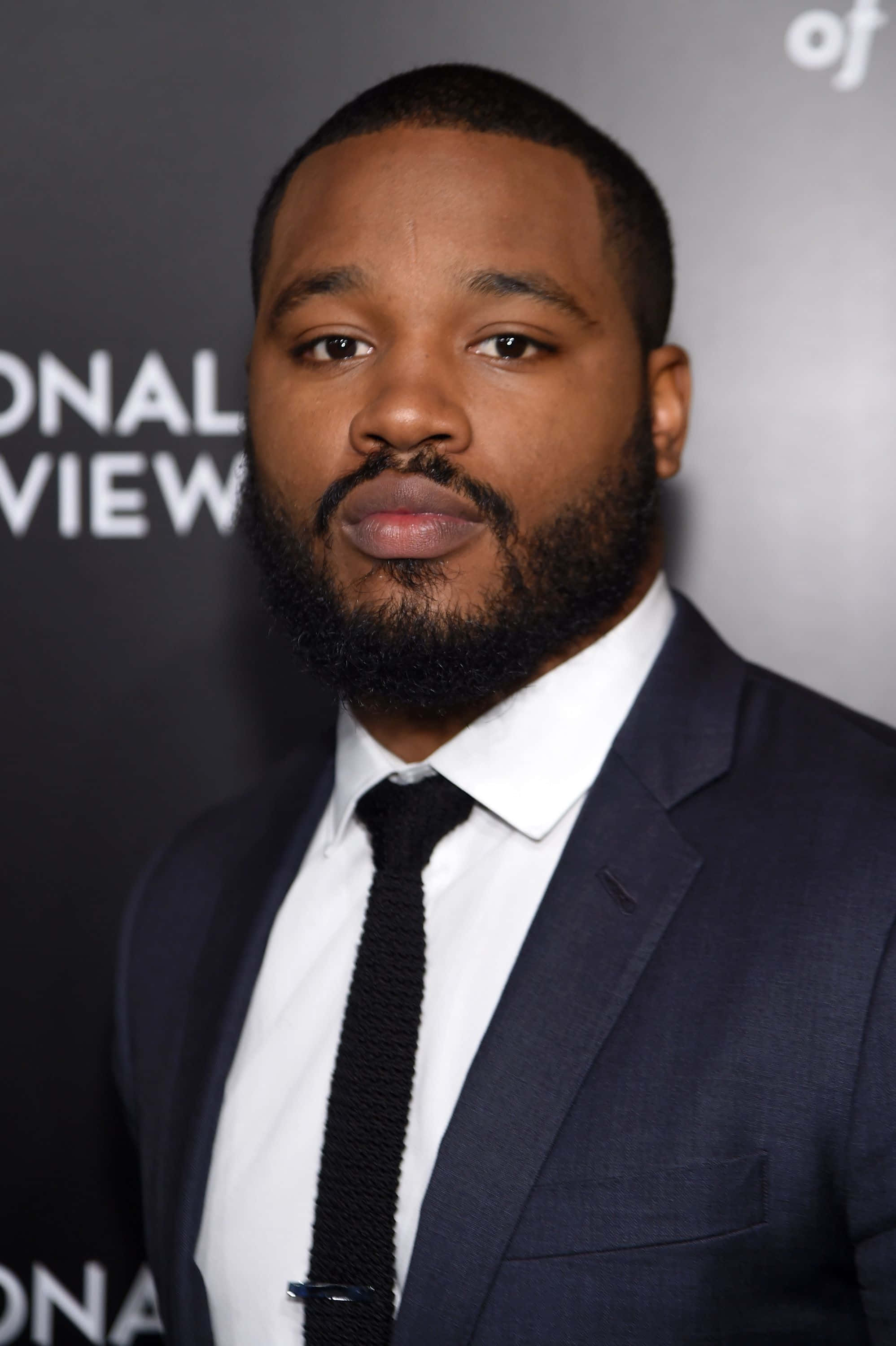 Ryan Coogler 桌布 壁紙 — 一位留鬍鬚的男子穿著深色西裝和黑色領帶，在品牌背景前的正式活動中擺姿勢。