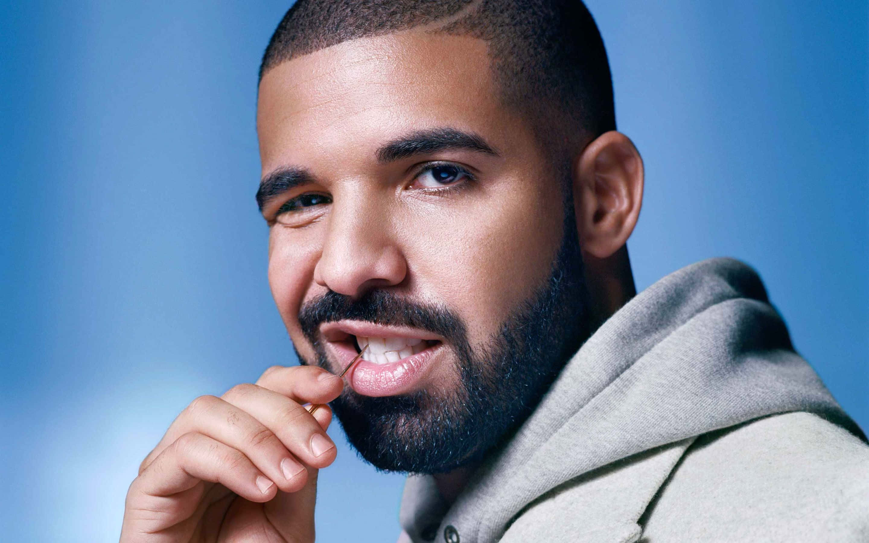 Drake 桌布 壁紙 — 一位留鬍鬚的男子穿著灰色連帽衫，在柔和的藍色背景前的特寫肖像。