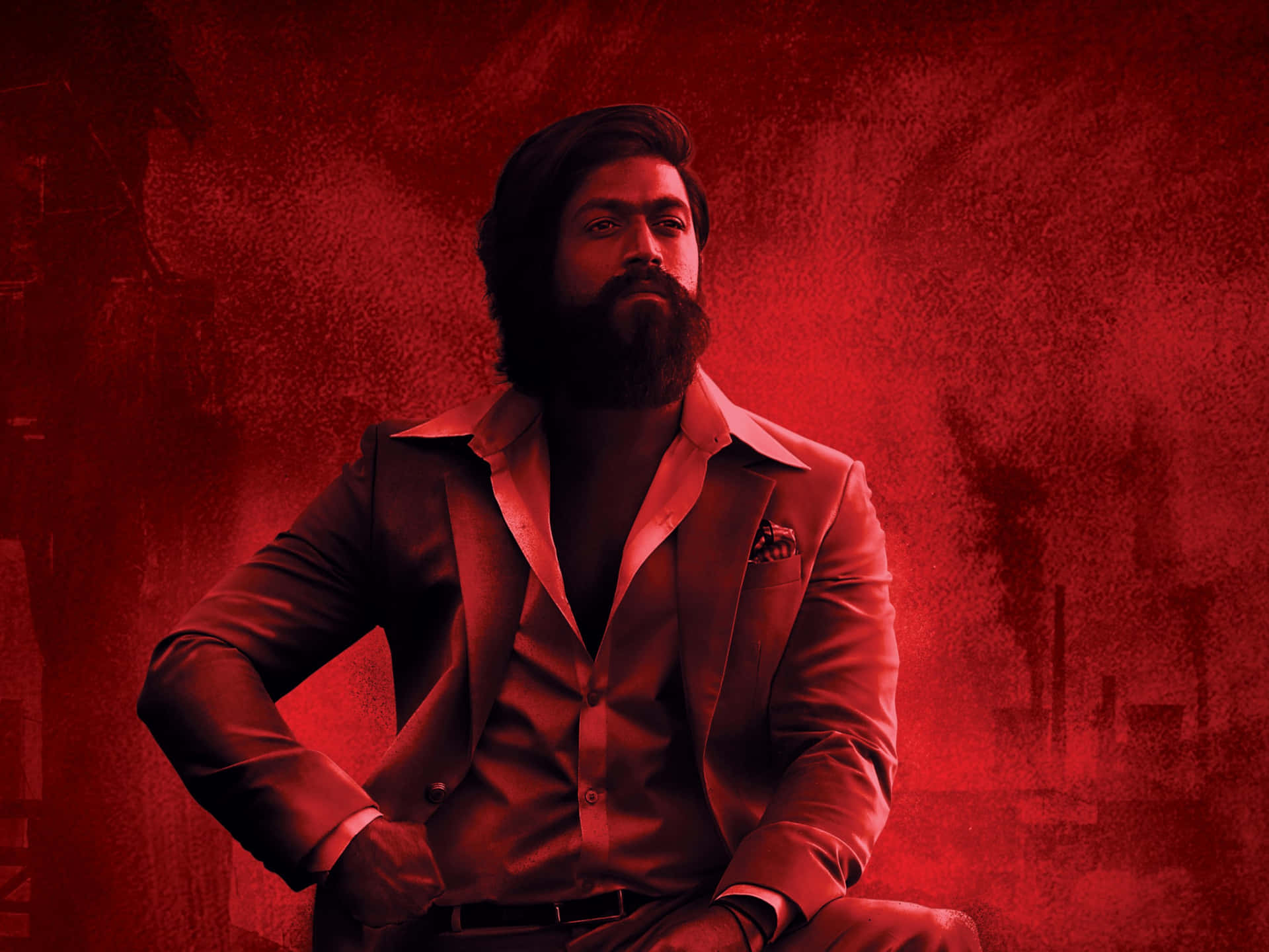 Kgf Chapter 2 桌布 壁紙 — 一位留鬍鬚的男子身穿俐落西裝，自信地坐在帶有工業剪影的紋理紅色背景前。
