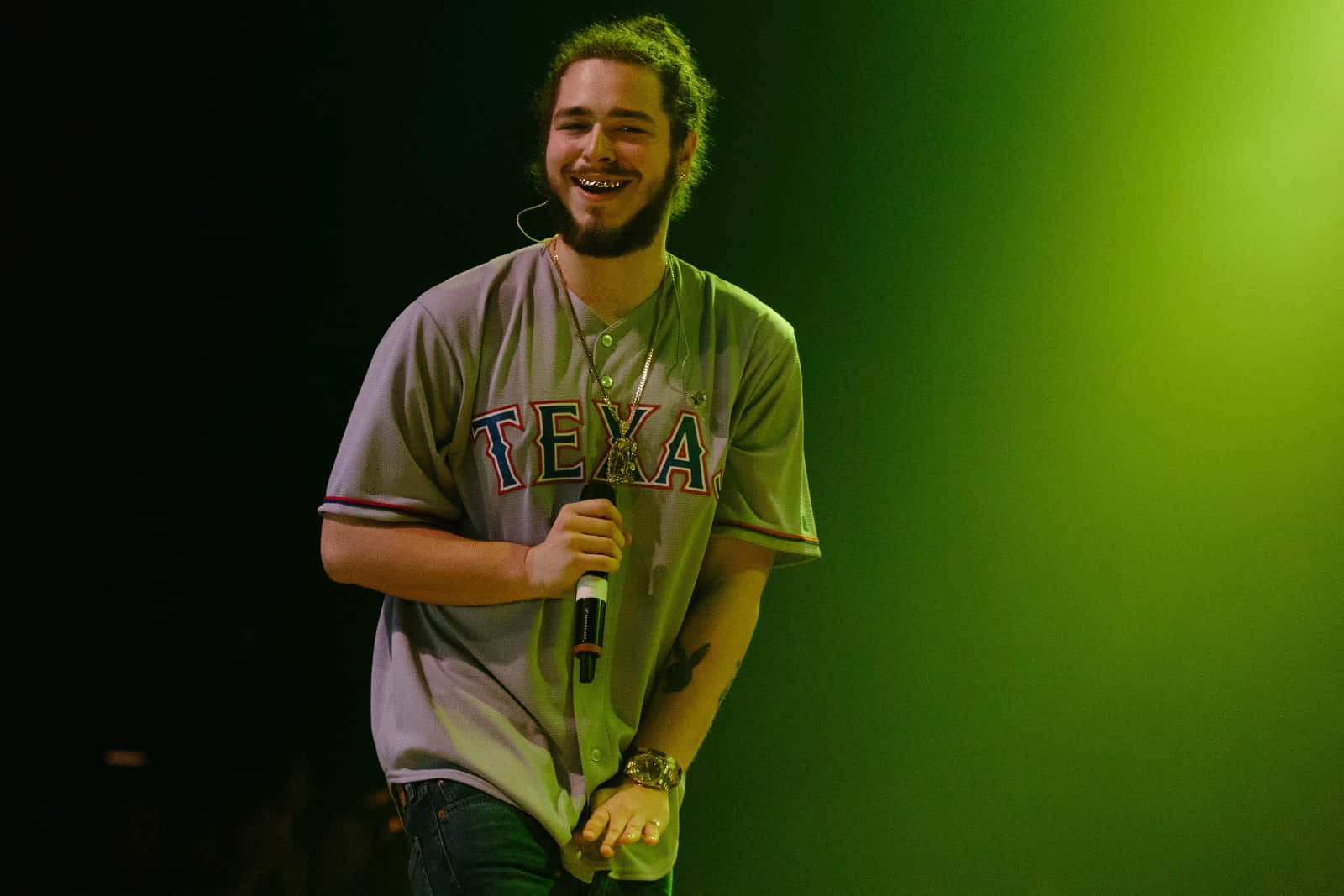 Post Malone 桌布 壁紙 — 一位留鬍鬚的男子身穿德州球衣，在綠色舞台燈光下微笑著拿著麥克風。