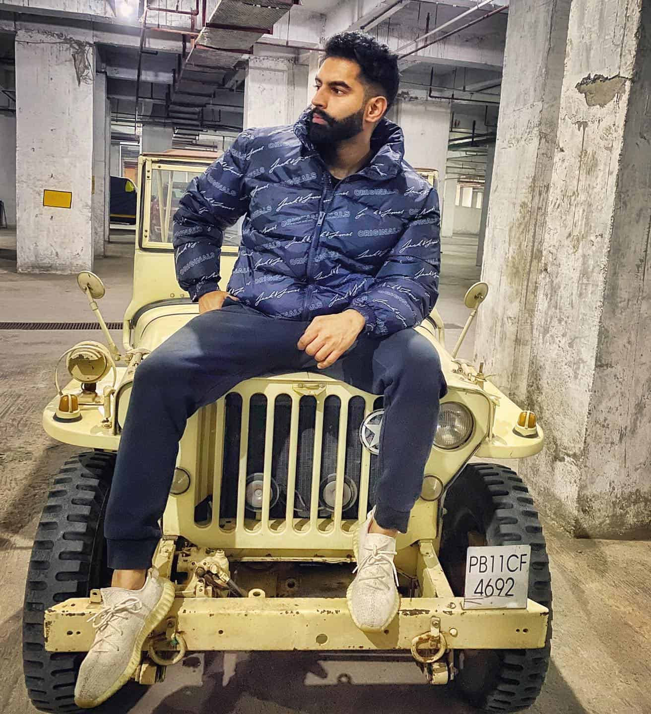 Indian 桌布 壁紙 — 一位留鬍鬚的男子身穿藍色圖案外套，自信地坐在一輛停在昏暗混凝土停車場的復古米色吉普車上。