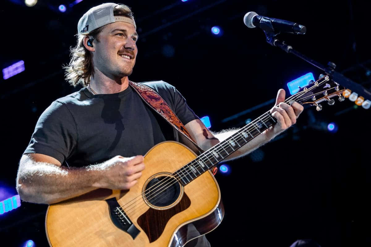 Morgan Wallen 桌布 壁紙 — 一位留鬍鬚的男音樂家面帶微笑，在現場音樂會表演期間於舞台燈光下彈奏木結他。