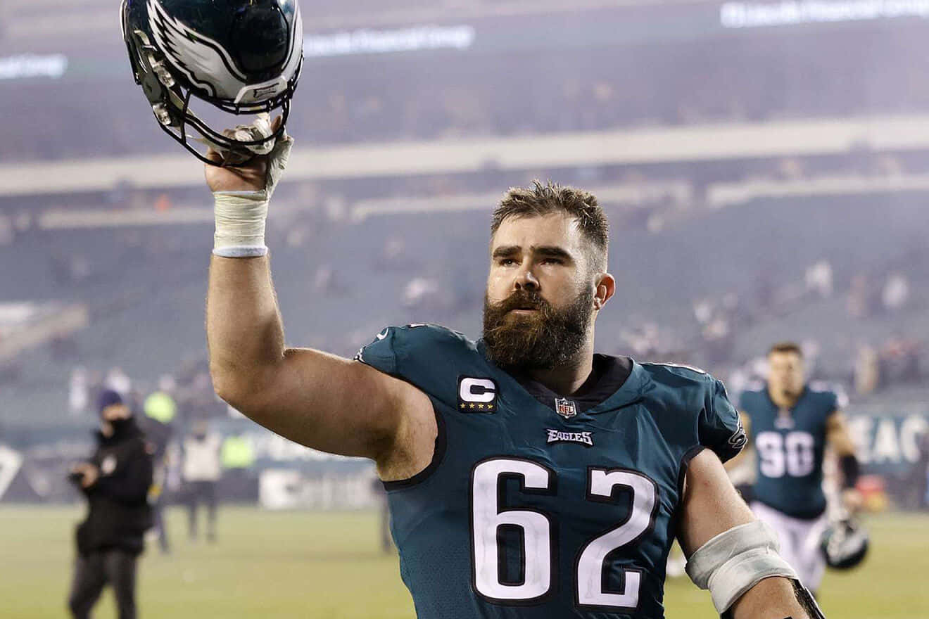 Jason Kelce 桌布 壁紙 — 一位留鬍鬚的費城老鷹隊美式足球員，身穿62號球衣，在夜晚的球場上高舉頭盔慶祝勝利。