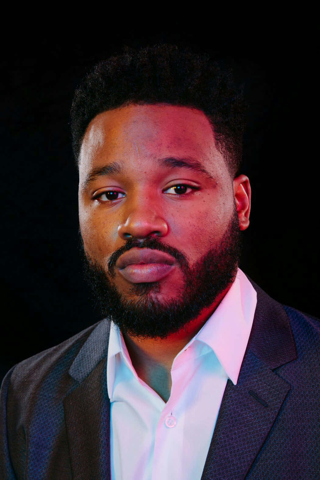 Ryan Coogler 桌布 壁紙 — 一位留鬍鬚短髮的男子，在黑色背景上戲劇性的紅藍燈光下穿著西裝和白襯衫。