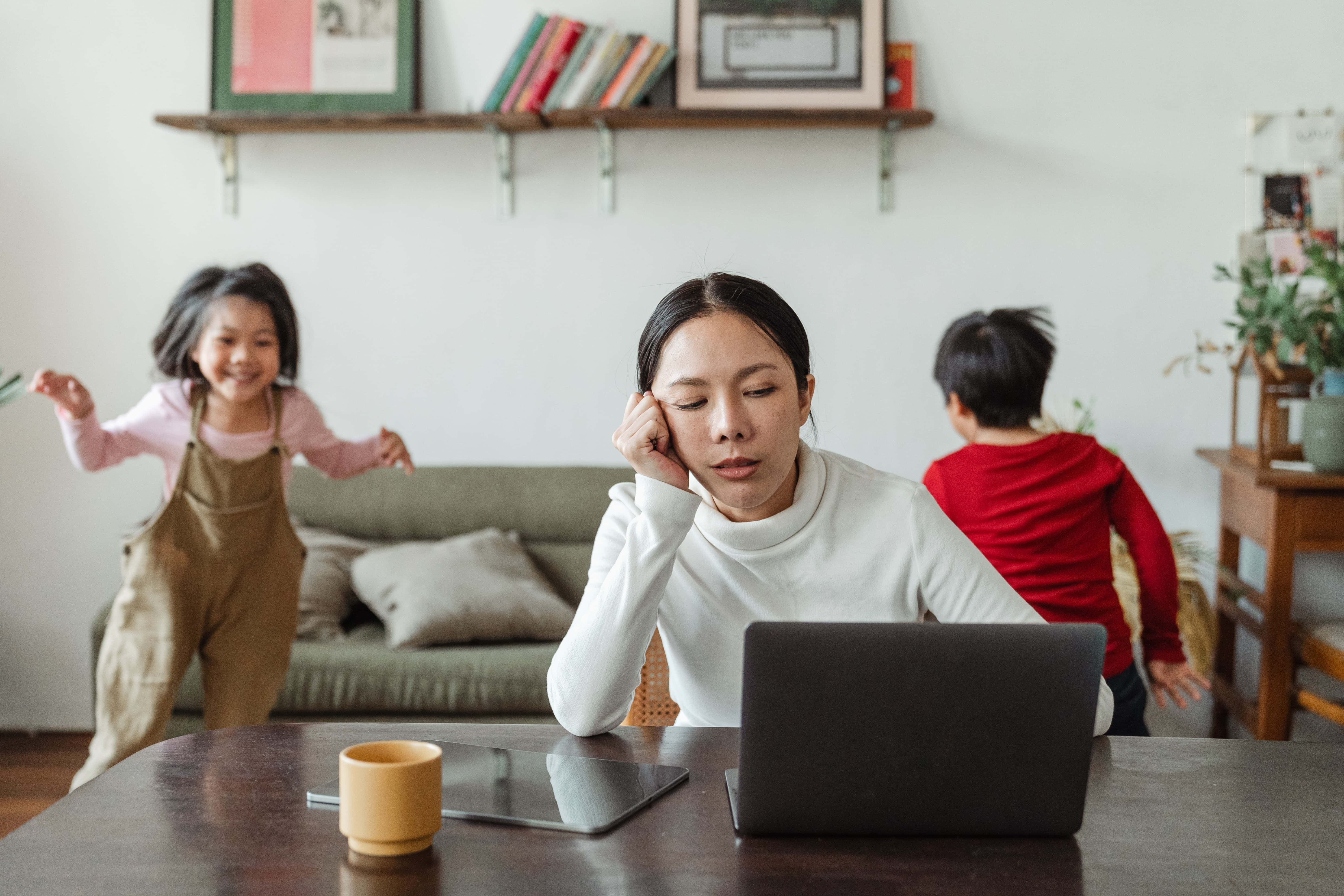 媽媽 桌布 壁紙 — 一位疲倦的女士在工作時用手托著頭，兩個孩子在她身後明亮的家中精力充沛地玩耍。